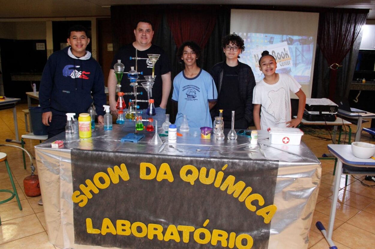 Na primeira fotografia há um grupo de estudantes apresentando o projeto do Clube de Ciências “Entre o passado e o presente, o que fica é a memória da gente”. A exposição conta com objetos antigos e decoração com fitas coloridas, promovendo uma reflexão sobre a memória. Na segunda imagem é possível ver um grupo de estudantes e um professor posando para a foto em um ambiente escolar. Eles estão atrás de uma mesa coberta com um tecido azul e verde, onde estão dispostos três baldes verdes rotulados como “Composteira” e um cesto com recipientes contendo composto. A terceira imagem mostra um grupo formado por quatro estudantes e um adulto posando atrás de uma mesa com diversos materiais de laboratório, como vidrarias, funis, tubos de ensaio e líquidos coloridos. À frente da mesa, há um painel com a frase “Show da Química Laboratório” em letras grandes e amarelas. O ambiente parece ser uma sala de aula com cortinas escuras ao fundo e um projetor exibindo uma tela com computador. A última imagem mostra uma sala de aula equipada como laboratório de ciências, com paredes azulejadas, bancadas de granito e modelos anatômicos expostos ao fundo. Diversos estudantes estão sentados ou conversando em grupos, enquanto alguns utilizam celulares ou notebooks. Um adulto, de camiseta azul, está sentado próximo à janela conversando com os alunos.