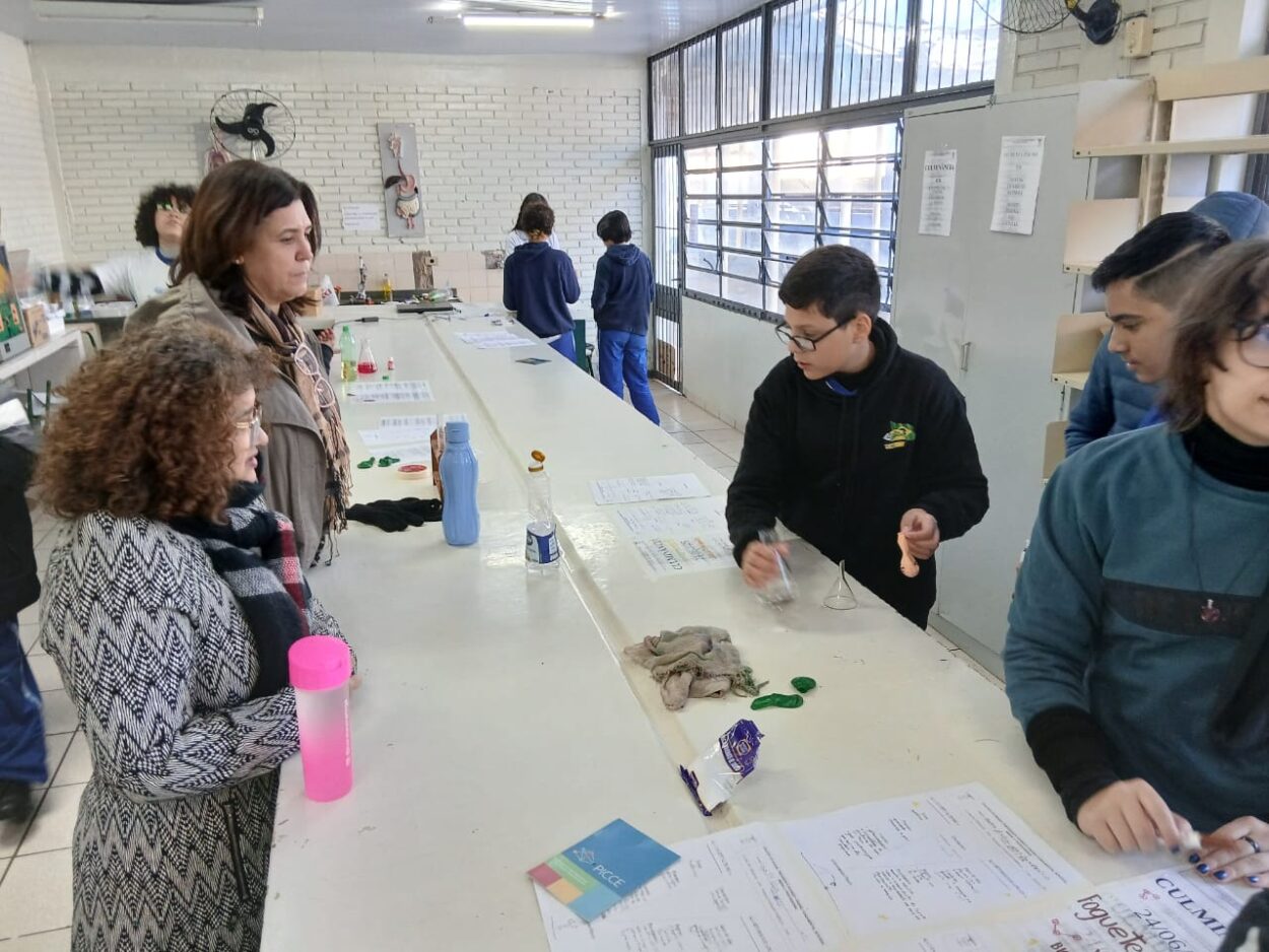 A imagem é uma galeria composta por seis fotografias, que retratam atividades desenvolvidas por diferentes grupos escolares ligados a Clubes de Ciências. A primeira foto tem um grupo de alunos sorrindo. O grupo está sentado em um ambiente interno decorado com plantas em prateleiras e cartazes. A segunda imagem mostra uma sala de informática com vários estudantes sentados em frente a computadores de mesa. Eles estão organizados em duas fileiras, cada uma voltada para o centro da sala, com monitores, teclados e fones de ouvido disponíveis. A maioria dos alunos parecem estar concentrados nas telas, digitando ou usando os fones. A sala tem iluminação fluorescente, paredes claras e alguns avisos afixados. Ao fundo, há ventiladores de parede e janelas com blocos de vidro para ventilação. O ambiente transmite uma atmosfera de estudo. A terceira fotografia tem alunos e visitantes interagindo em uma bancada comprida, realizando atividades práticas e experimentos. Há papéis com explicações e materiais sobre a mesa, indicando uma exposição interativa. A quarta foto mostra várias pessoas observando atentamente uma exposição de trabalhos em um espaço decorado com tecido verde. Na quinta foto tem um grupo de alunos apresentando um projeto do Clube de Ciências em uma sala decorada com folhas verdes penduradas. Na frente da mesa, há cartazes escritos e materiais como garrafas PET e experimentos. Na última foto há um grupo de estudantes, alguns vestidos com roupas típicas de festa junina, participando de uma atividade que envolve códigos QR. Uma das alunas segura um cartaz com o título “Quiz da ciência”, sugerindo uma abordagem lúdica para aprender conteúdos científicos. 