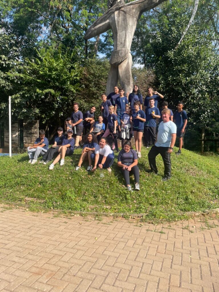 Na primeira imagem há uma sala de aula onde um grupo de estudantes está participando de uma atividade. Alguns estão sentados ao redor de uma mesa utilizando notebooks, enquanto um aluno em pé apresenta algo projetado em um quadro branco, apontando para um gráfico exibido na tela. O ambiente tem paredes claras, iluminação fluorescente e móveis escolares, como cadeiras e armários. Na segunda fotografia tem um grupo de estudantes reunidos em um ambiente escolar ao ar livre, em frente a um jardim vertical com diversas plantas verdes. Eles estão distribuídos em pé e agachados, posando para a foto. O local tem piso de tijolos, árvores, telhados de cerâmica e áreas cobertas ao fundo. A terceira imagem mostra um grupo de estudantes e um adulto reunidos em um espaço ao ar livre, posando para uma foto em frente a uma grande escultura de uma figura humana com os braços abertos. O grupo está distribuído sobre um pequeno morro coberto de grama, alguns sentados e outros em pé, enquanto ao fundo há muitas árvores, formando um cenário verde e natural. Na quarta imagem há um grupo de estudantes e dois adultos posando para uma foto em frente a uma placa de madeira com a palavra “Meliponário” e o desenho de uma abelha sorridente. Eles estão em um espaço externo com bastante vegetação, incluindo árvores altas e um caminho de pedras à esquerda. Ao fundo, vê-se um prédio de tijolos com telhado de cerâmica. Na quinta imagem tem um grupo de estudantes e alguns adultos reunidos para uma foto em um ambiente escolar externo. Eles estão em frente a uma parede branca com detalhes verdes e uma janela comprida com grades. O grupo está posicionado em duas fileiras, com alguns agachados na frente e outros em pé atrás, todos sorrindo e olhando para a câmera. A última fotografia mostra um grupo grande de pessoas, formado por adolescentes e um adulto, posando para uma foto em uma sala. Eles estão organizados em duas fileiras: alguns sentados ou agachados na frente e outros em pé atrás. A sala tem paredes claras, ventiladores fixados na parede e um quadro branco ao fundo. Há cartazes e avisos na parede. À esquerda, há uma mesa com uma toalha vermelha e alguns objetos decorativos para um evento escolar.