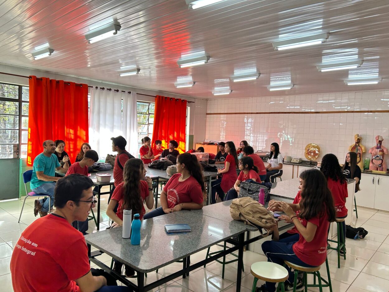Na primeira fotografia há um grupo de estudantes apresentando o projeto do Clube de Ciências “Entre o passado e o presente, o que fica é a memória da gente”. A exposição conta com objetos antigos e decoração com fitas coloridas, promovendo uma reflexão sobre a memória. Na segunda imagem é possível ver um grupo de estudantes e um professor posando para a foto em um ambiente escolar. Eles estão atrás de uma mesa coberta com um tecido azul e verde, onde estão dispostos três baldes verdes rotulados como “Composteira” e um cesto com recipientes contendo composto. A terceira imagem mostra um grupo formado por quatro estudantes e um adulto posando atrás de uma mesa com diversos materiais de laboratório, como vidrarias, funis, tubos de ensaio e líquidos coloridos. À frente da mesa, há um painel com a frase “Show da Química Laboratório” em letras grandes e amarelas. O ambiente parece ser uma sala de aula com cortinas escuras ao fundo e um projetor exibindo uma tela com computador. A última imagem mostra uma sala de aula equipada como laboratório de ciências, com paredes azulejadas, bancadas de granito e modelos anatômicos expostos ao fundo. Diversos estudantes estão sentados ou conversando em grupos, enquanto alguns utilizam celulares ou notebooks. Um adulto, de camiseta azul, está sentado próximo à janela conversando com os alunos.