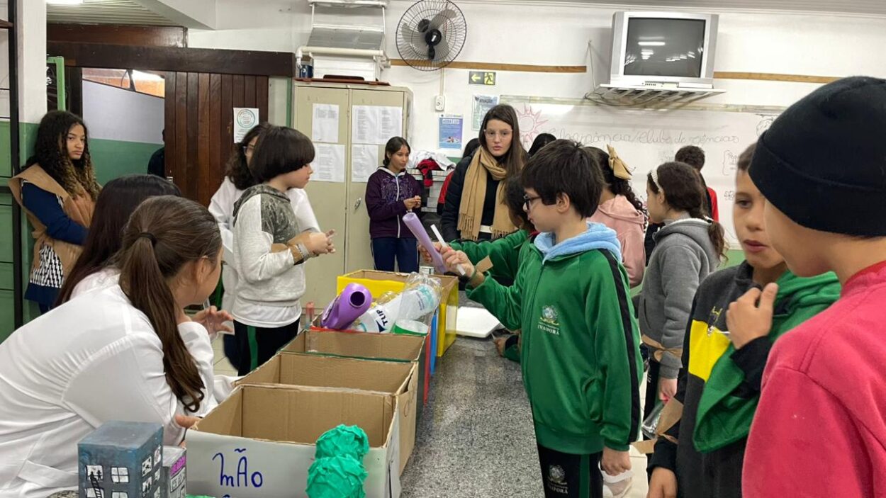 A galeria é composta por cinco fotografias, retratando diferentes momentos de atividades escolares desenvolvidas pelos Clubes de Ciências. A primeira foto expõe um grupo de estudantes e um professor posicionados atrás de três composteiras verdes com etiquetas, em um projeto que aborda o reaproveitamento de resíduos orgânicos. A segunda foto mostra alunos e professores posando ao redor de uma bancada com frascos, reagentes e uma grande bacia. A cena remete a um experimento de ciências em andamento, demonstrando práticas laboratoriais e espírito de equipe. Na terceira fotografia há um grupo de estudantes apresentando o projeto do Clube de Ciências “Entre o passado e o presente, o que fica é a memória da gente”. A exposição conta com objetos antigos e decoração com fitas coloridas, promovendo uma reflexão sobre a memória. A quarta foto mostra os alunos participando de uma atividade interativa sobre reciclagem, utilizando caixas organizadas por tipo de resíduo (plástico, papel, etc.). Crianças observam e manuseiam objetos, aprendendo sobre o descarte correto de materiais. Na última foto há dois alunos que apresentam um painel, ao lado de caixas de coleta seletiva. Uma aluna observa o espaço, que contém cartazes e materiais ligados à preservação ambiental.