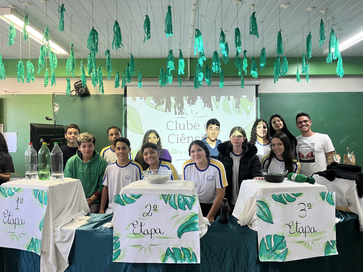 A imagem é uma galeria composta por seis fotografias, que retratam atividades desenvolvidas por diferentes grupos escolares ligados a Clubes de Ciências. A primeira foto tem um grupo de alunos sorrindo. O grupo está sentado em um ambiente interno decorado com plantas em prateleiras e cartazes. A segunda imagem mostra uma sala de informática com vários estudantes sentados em frente a computadores de mesa. Eles estão organizados em duas fileiras, cada uma voltada para o centro da sala, com monitores, teclados e fones de ouvido disponíveis. A maioria dos alunos parecem estar concentrados nas telas, digitando ou usando os fones. A sala tem iluminação fluorescente, paredes claras e alguns avisos afixados. Ao fundo, há ventiladores de parede e janelas com blocos de vidro para ventilação. O ambiente transmite uma atmosfera de estudo. A terceira fotografia tem alunos e visitantes interagindo em uma bancada comprida, realizando atividades práticas e experimentos. Há papéis com explicações e materiais sobre a mesa, indicando uma exposição interativa. A quarta foto mostra várias pessoas observando atentamente uma exposição de trabalhos em um espaço decorado com tecido verde. Na quinta foto tem um grupo de alunos apresentando um projeto do Clube de Ciências em uma sala decorada com folhas verdes penduradas. Na frente da mesa, há cartazes escritos e materiais como garrafas PET e experimentos. Na última foto há um grupo de estudantes, alguns vestidos com roupas típicas de festa junina, participando de uma atividade que envolve códigos QR. Uma das alunas segura um cartaz com o título “Quiz da ciência”, sugerindo uma abordagem lúdica para aprender conteúdos científicos. 