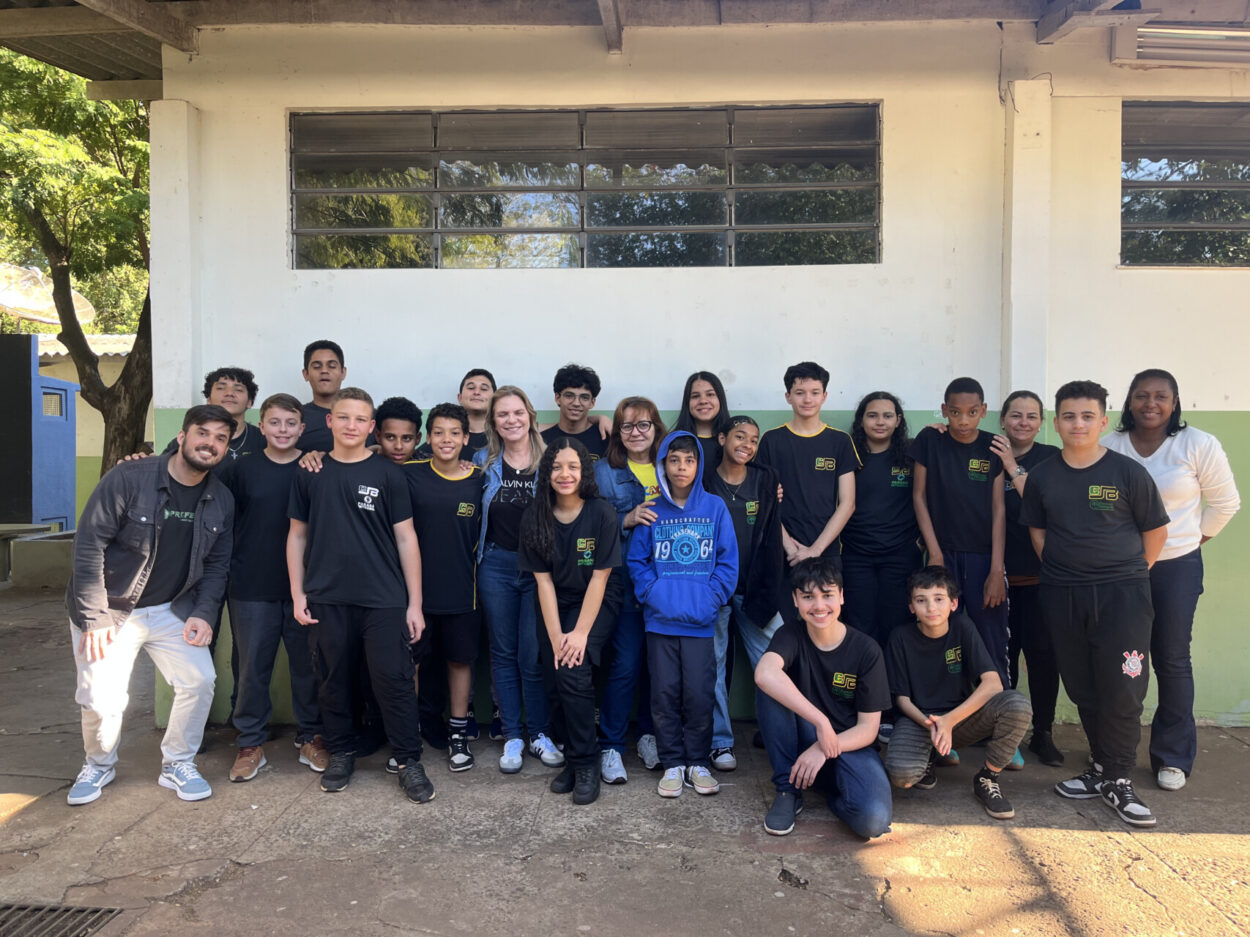 Na primeira imagem há uma sala de aula onde um grupo de estudantes está participando de uma atividade. Alguns estão sentados ao redor de uma mesa utilizando notebooks, enquanto um aluno em pé apresenta algo projetado em um quadro branco, apontando para um gráfico exibido na tela. O ambiente tem paredes claras, iluminação fluorescente e móveis escolares, como cadeiras e armários. Na segunda fotografia tem um grupo de estudantes reunidos em um ambiente escolar ao ar livre, em frente a um jardim vertical com diversas plantas verdes. Eles estão distribuídos em pé e agachados, posando para a foto. O local tem piso de tijolos, árvores, telhados de cerâmica e áreas cobertas ao fundo. A terceira imagem mostra um grupo de estudantes e um adulto reunidos em um espaço ao ar livre, posando para uma foto em frente a uma grande escultura de uma figura humana com os braços abertos. O grupo está distribuído sobre um pequeno morro coberto de grama, alguns sentados e outros em pé, enquanto ao fundo há muitas árvores, formando um cenário verde e natural. Na quarta imagem há um grupo de estudantes e dois adultos posando para uma foto em frente a uma placa de madeira com a palavra “Meliponário” e o desenho de uma abelha sorridente. Eles estão em um espaço externo com bastante vegetação, incluindo árvores altas e um caminho de pedras à esquerda. Ao fundo, vê-se um prédio de tijolos com telhado de cerâmica. Na quinta imagem tem um grupo de estudantes e alguns adultos reunidos para uma foto em um ambiente escolar externo. Eles estão em frente a uma parede branca com detalhes verdes e uma janela comprida com grades. O grupo está posicionado em duas fileiras, com alguns agachados na frente e outros em pé atrás, todos sorrindo e olhando para a câmera. A última fotografia mostra um grupo grande de pessoas, formado por adolescentes e um adulto, posando para uma foto em uma sala. Eles estão organizados em duas fileiras: alguns sentados ou agachados na frente e outros em pé atrás. A sala tem paredes claras, ventiladores fixados na parede e um quadro branco ao fundo. Há cartazes e avisos na parede. À esquerda, há uma mesa com uma toalha vermelha e alguns objetos decorativos para um evento escolar.
