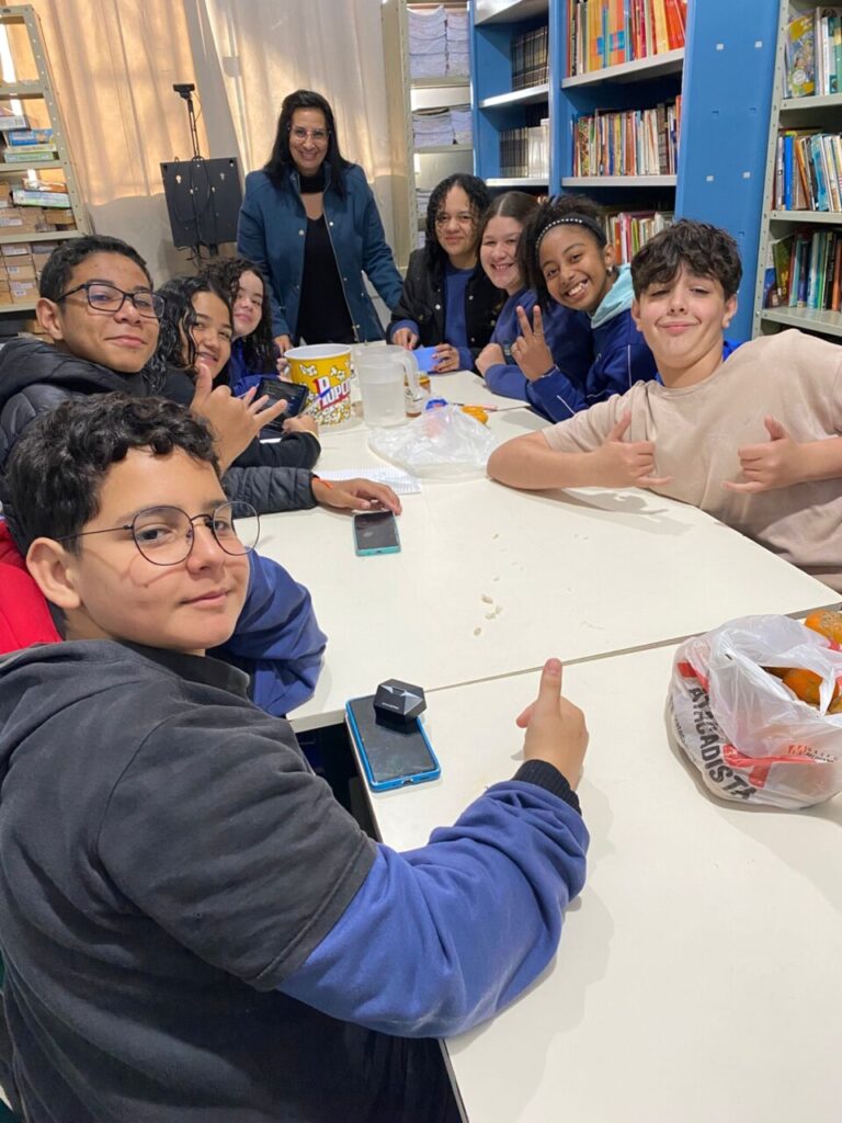A imagem mostra um grupo de nove estudantes reunidos ao redor de uma mesa branca em uma biblioteca escolar, acompanhados por uma professora em pé ao fundo. Todos parecem felizes, sorrindo e fazendo gestos descontraídos com as mãos. A mesa está repleta de itens como um balde de pipoca, jarra com água, celular, pote de mel, sacola com laranjas e cadernos. Ao fundo, há prateleiras cheias de livros, reforçando o ambiente educativo. A cena transmite um clima de amizade, aprendizado e acolhimento.