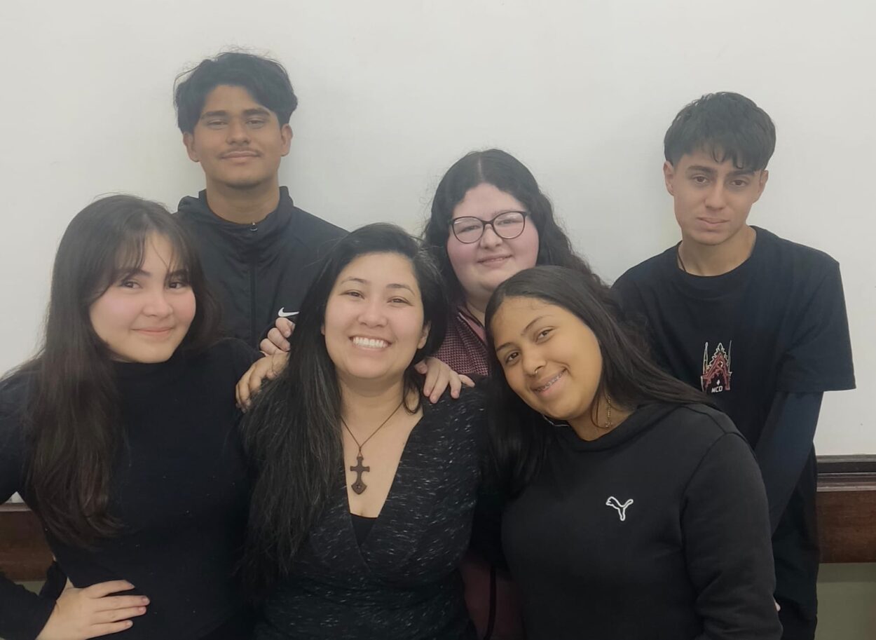 A imagem mostra seis pessoas reunidas, em ambiente interno com parede branca ao fundo. Todos estão próximos, sorrindo ou olhando para a câmera. Na frente, quatro pessoas aparecem lado a lado, e atrás delas, duas outras completam o grupo. A cena transmite amizade e acolhimento, com destaque para as expressões felizes e o clima de união.