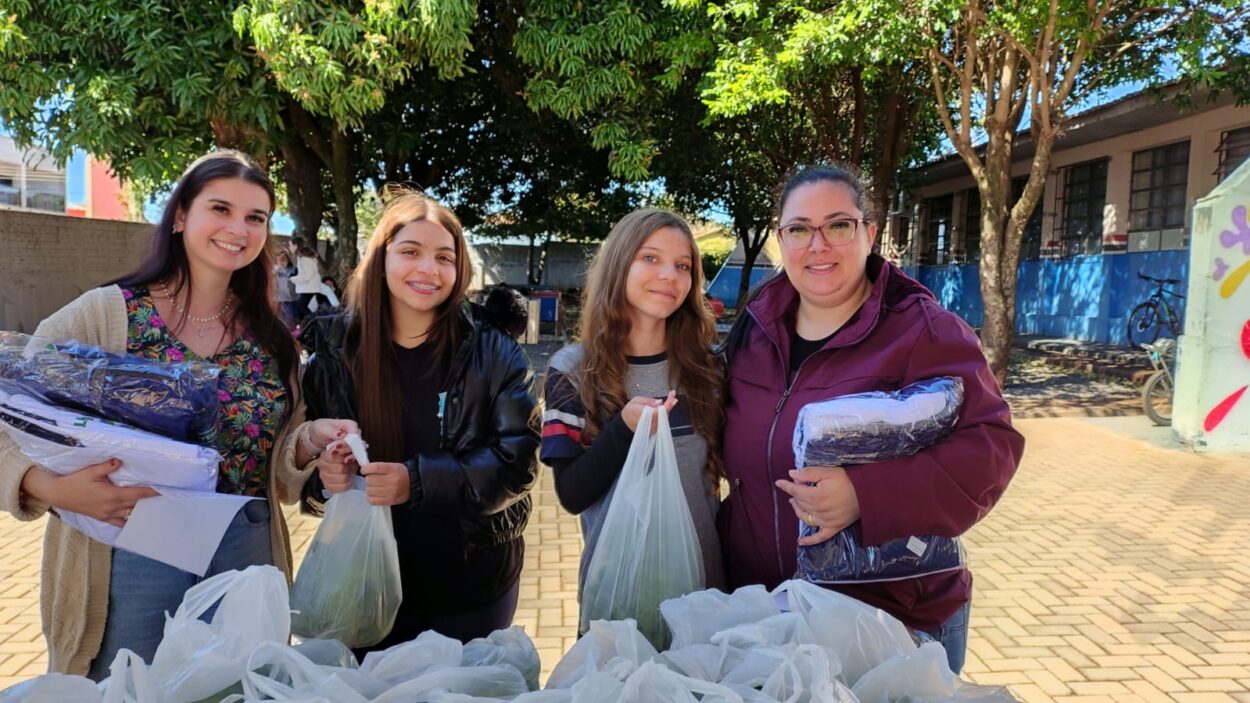 A imagem mostra quatro mulheres sorrindo, em um ambiente externo com árvores ao fundo e parte de um prédio escolar visível à direita. Elas estão posicionadas atrás de uma mesa coberta por diversas sacolas plásticas brancas. Cada uma segura sacolas semelhantes ou pacotes contendo uniformes escolares. À esquerda, uma das mulheres segura um maço de roupas dobradas com etiquetas, enquanto outra segura uma sacola plástica com as duas mãos. Ao fundo, outras pessoas e bicicletas podem ser vistas, reforçando o clima comunitário e escolar da cena. A iluminação é natural e o clima parece estar ameno.