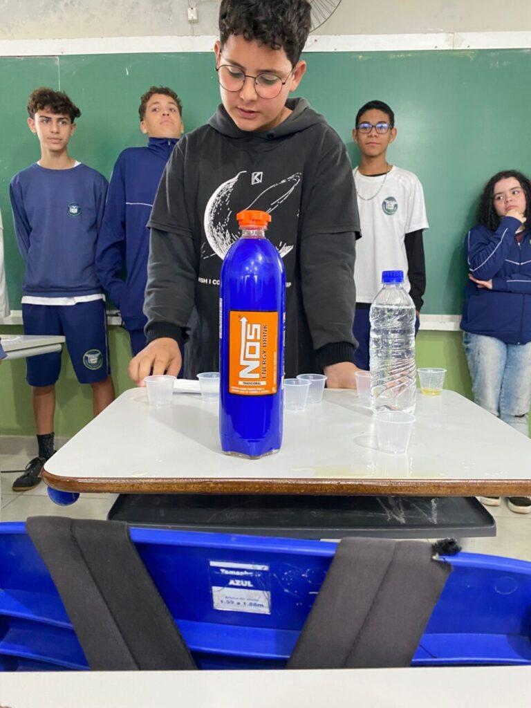 A galeria de fotos mostra momentos da apresentação e degustação do energético produzido pelo clube de ciências Ciência na Mesa. As imagens mostram estudantes na sala de aula com as mesas organizadas para oferecer o energético em copo descartáveis. Todos parecem felizes, sorrindo e curiosos com a proposta de bebida alternativa