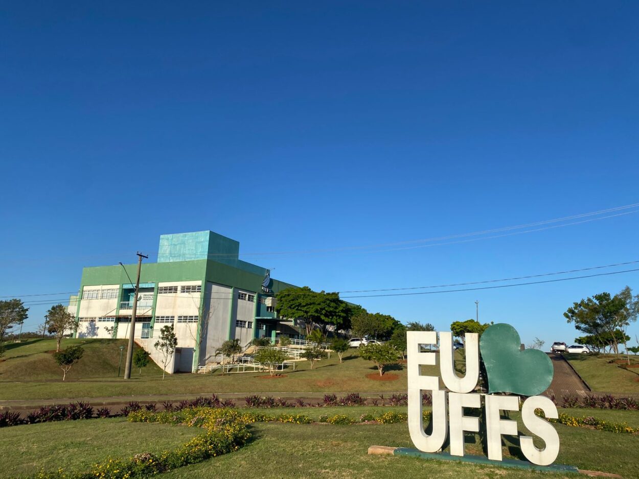 A foto mostra a fachada de um prédio da Universidade Federal da Fronteira Sul (UFFS), no campus de Realeza. O edifício é de dois andares, pintado em tons de verde e branco, com janelas retangulares alinhadas. Em frente, há um gramado bem cuidado com canteiros de flores. No lado direito, destaca-se um letreiro grande em branco escrito “EU ♥ UFFS”, onde o coração é representado por uma forma verde. Ao fundo, aparecem árvores, carros estacionados e um céu azul claro, sem nuvens.
