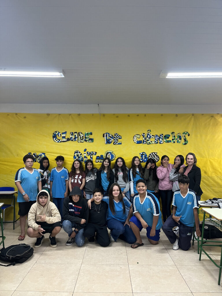 A primeira imagem mostra um grupo de estudantes e uma professora em uma sala de aula em frente ao mural do clube “No Ritmo do Conhecimento”. As crianças estão sorrindo e posicionadas ao redor de uma mesa com cartazes coloridos e materiais escolares espalhados. A segunda imagem mostra um grupo de estudantes posando para uma foto dentro de uma sala de aula. O grupo é formado por 14 alunos e uma adulta, a professora. Eles estão dispostos em duas fileiras: a primeira sentada e a segunda em pé. Atrás deles há um mural com cartazes e trabalhos escolares expostos na parede. A terceira foto mostra um grupo de estudantes e uma professora reunidos em uma sala de aula. Eles estão posando para uma foto em frente a um quadro verde, no qual está escrito “Clube de Ciências”. No ambiente, é possível ver carteiras na frente, uma televisão ligada exibindo a palavra “Conceitos Botânicos” e uma janela com grades à esquerda. A quarta fotografia é do Clube Jovens Cientistas Kennedy. A imagem mostra um grupo grande de estudantes posando para a foto em um pátio externo, com árvores de folhagem vermelha ao fundo e um muro branco atrás deles. Algumas alunas estão agachadas na frente, junto com duas mulheres adultas, enquanto o restante da turma permanece em pé. Um homem adulto está à esquerda do grupo. A quinta fotografia mostra um grupo de estudantes reunidos em uma sala de aula, participando do Clube de Ciência. Eles vestem camisetas verdes do colégio e estão organizados ao redor de uma mesa de laboratório. Ao fundo, há uma tela que apresenta o tema do projeto: “Plant Scientists Club: Descobrindo alternativas no combate de plantas daninhas a partir do estudo de bioinsumos”. Na sexta imagem, um grupo de estudantes e professores está reunido em um espaço externo, em frente a árvores e cercas metálicas. Eles posam sorridentes para a foto. Na frente, duas mulheres seguram uma bandeira verde e branca com o desenho de uma capivara e os dizeres “CAPCIÊNCIA – Clube de Ciências”.