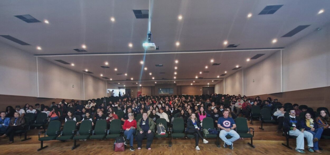 A imagem mostra um auditório grande, bem iluminado, com muitas pessoas sentadas em cadeiras verdes organizadas em fileiras. O chão é de madeira clara, o teto branco e o ambiente tem iluminação embutida com várias luzes circulares distribuídas em linhas. As cadeiras estão praticamente todas ocupadas, principalmente no centro e no fundo.