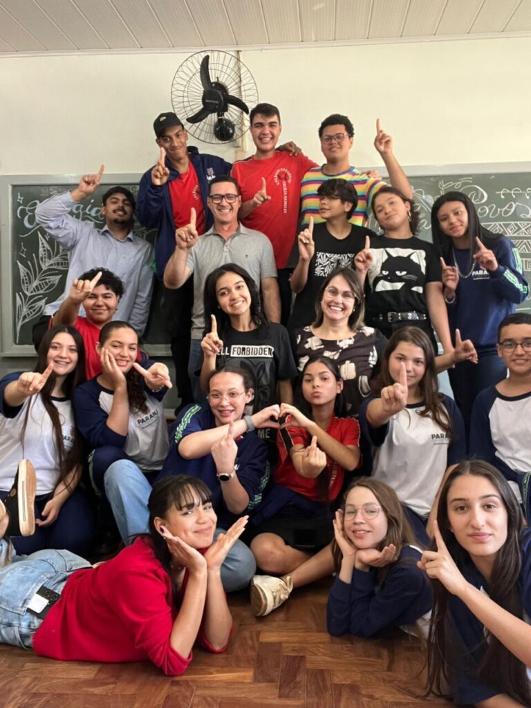 A imagem mostra um grupo alegre de estudantes e professores posando para uma foto em sala de aula. Há cerca de 25 pessoas, entre jovens e adultos, organizadas em três fileiras: alguns estão em pé ao fundo, outros agachados ou sentados no meio, e um grupo está sentado ou deitado na frente, no chão. Todos estão sorrindo e fazendo poses animadas com os dedos apontando para cima, formando corações com as mãos ou apoiando o rosto com as mãos em sinal de carinho e alegria. Há também um quadro ao fundo com uma bela decoração de letras cursivas e desenhos, indicando uma ocasião especial, como uma festa.