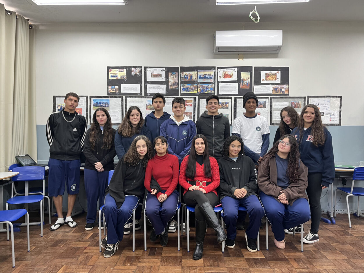 A primeira imagem mostra um grupo de estudantes e uma professora em uma sala de aula em frente ao mural do clube “No Ritmo do Conhecimento”. As crianças estão sorrindo e posicionadas ao redor de uma mesa com cartazes coloridos e materiais escolares espalhados. A segunda imagem mostra um grupo de estudantes posando para uma foto dentro de uma sala de aula. O grupo é formado por 14 alunos e uma adulta, a professora. Eles estão dispostos em duas fileiras: a primeira sentada e a segunda em pé. Atrás deles há um mural com cartazes e trabalhos escolares expostos na parede. A terceira foto mostra um grupo de estudantes e uma professora reunidos em uma sala de aula. Eles estão posando para uma foto em frente a um quadro verde, no qual está escrito “Clube de Ciências”. No ambiente, é possível ver carteiras na frente, uma televisão ligada exibindo a palavra “Conceitos Botânicos” e uma janela com grades à esquerda. A quarta fotografia é do Clube Jovens Cientistas Kennedy. A imagem mostra um grupo grande de estudantes posando para a foto em um pátio externo, com árvores de folhagem vermelha ao fundo e um muro branco atrás deles. Algumas alunas estão agachadas na frente, junto com duas mulheres adultas, enquanto o restante da turma permanece em pé. Um homem adulto está à esquerda do grupo. A quinta fotografia mostra um grupo de estudantes reunidos em uma sala de aula, participando do Clube de Ciência. Eles vestem camisetas verdes do colégio e estão organizados ao redor de uma mesa de laboratório. Ao fundo, há uma tela que apresenta o tema do projeto: “Plant Scientists Club: Descobrindo alternativas no combate de plantas daninhas a partir do estudo de bioinsumos”. Na sexta imagem, um grupo de estudantes e professores está reunido em um espaço externo, em frente a árvores e cercas metálicas. Eles posam sorridentes para a foto. Na frente, duas mulheres seguram uma bandeira verde e branca com o desenho de uma capivara e os dizeres “CAPCIÊNCIA – Clube de Ciências”.
