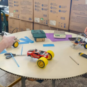 Dois alunos em uma sala de aula trabalhando em um projeto de robótica. Eles estão em pé em volta de uma mesa redonda branca, que possui sobre ela dois carrinhos de controle remoto ou protótipos de veículos. Um deles é vermelho com rodas amarelas, já montado e com aparência de brinquedo pronto; o outro está em processo de montagem, com fios, componentes eletrônicos e estrutura exposta. Na mesa também há ferramentas e materiais.