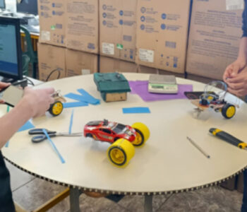 Dois alunos em uma sala de aula trabalhando em um projeto de robótica. Eles estão em pé em volta de uma mesa redonda branca, que possui sobre ela dois carrinhos de controle remoto ou protótipos de veículos. Um deles é vermelho com rodas amarelas, já montado e com aparência de brinquedo pronto; o outro está em processo de montagem, com fios, componentes eletrônicos e estrutura exposta. Na mesa também há ferramentas e materiais.