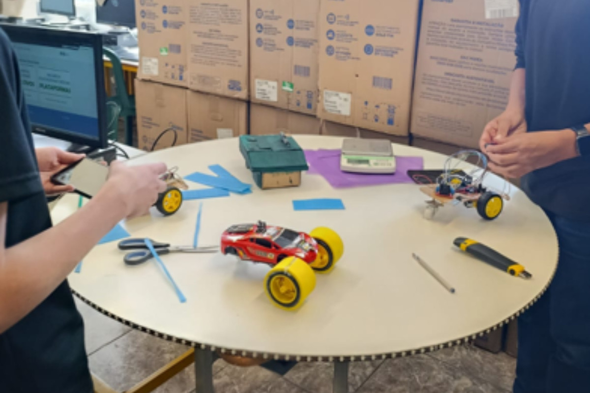 Dois alunos em uma sala de aula trabalhando em um projeto de robótica. Eles estão em pé em volta de uma mesa redonda branca, que possui sobre ela dois carrinhos de controle remoto ou protótipos de veículos. Um deles é vermelho com rodas amarelas, já montado e com aparência de brinquedo pronto; o outro está em processo de montagem, com fios, componentes eletrônicos e estrutura exposta. Na mesa também há ferramentas e materiais.