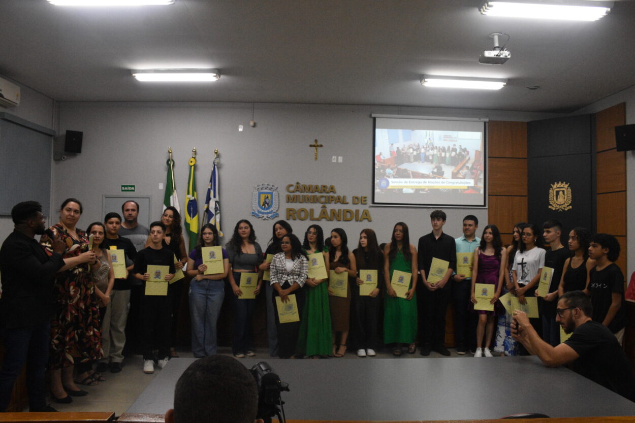 A foto mostra um grupo grande de pessoas reunidas em frente à parede da Câmara Municipal de Rolândia. Todos estão de pé, lado a lado, segurando certificados amarelos. Alguns usam roupas formais, outros mais casuais. Atrás deles há bandeiras. No alto da parede, vê-se uma cruz e o brasão da câmara. À direita, há um telão com uma imagem projetada mostrando a mesma cerimônia.