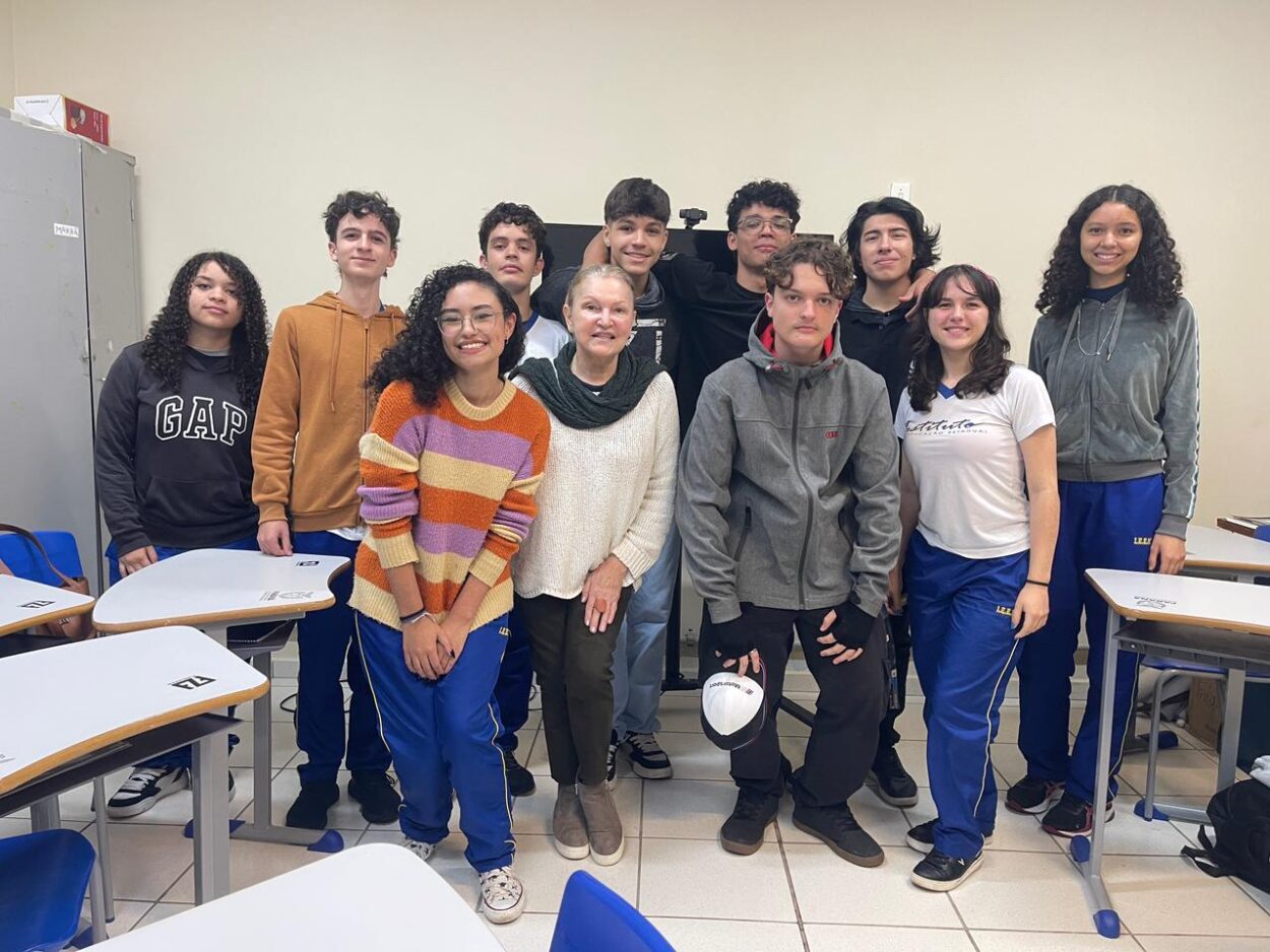 A foto é do clube de ciências ‘Cienteens’ do Colégio Instituto de Educação Estadual de Maringá. Na imagem, vemos um grupo de onze pessoas posando dentro de uma sala de aula. A maioria são jovens estudantes, acompanhados por uma mulher adulta, que é a professora, posicionada no centro da foto. Eles estão sorridentes e de pé. Ao fundo, a parede é clara e há um armário metálico do lado esquerdo. À frente, aparecem mesas e cadeiras brancas com detalhes em madeira, típicas de sala de aula.