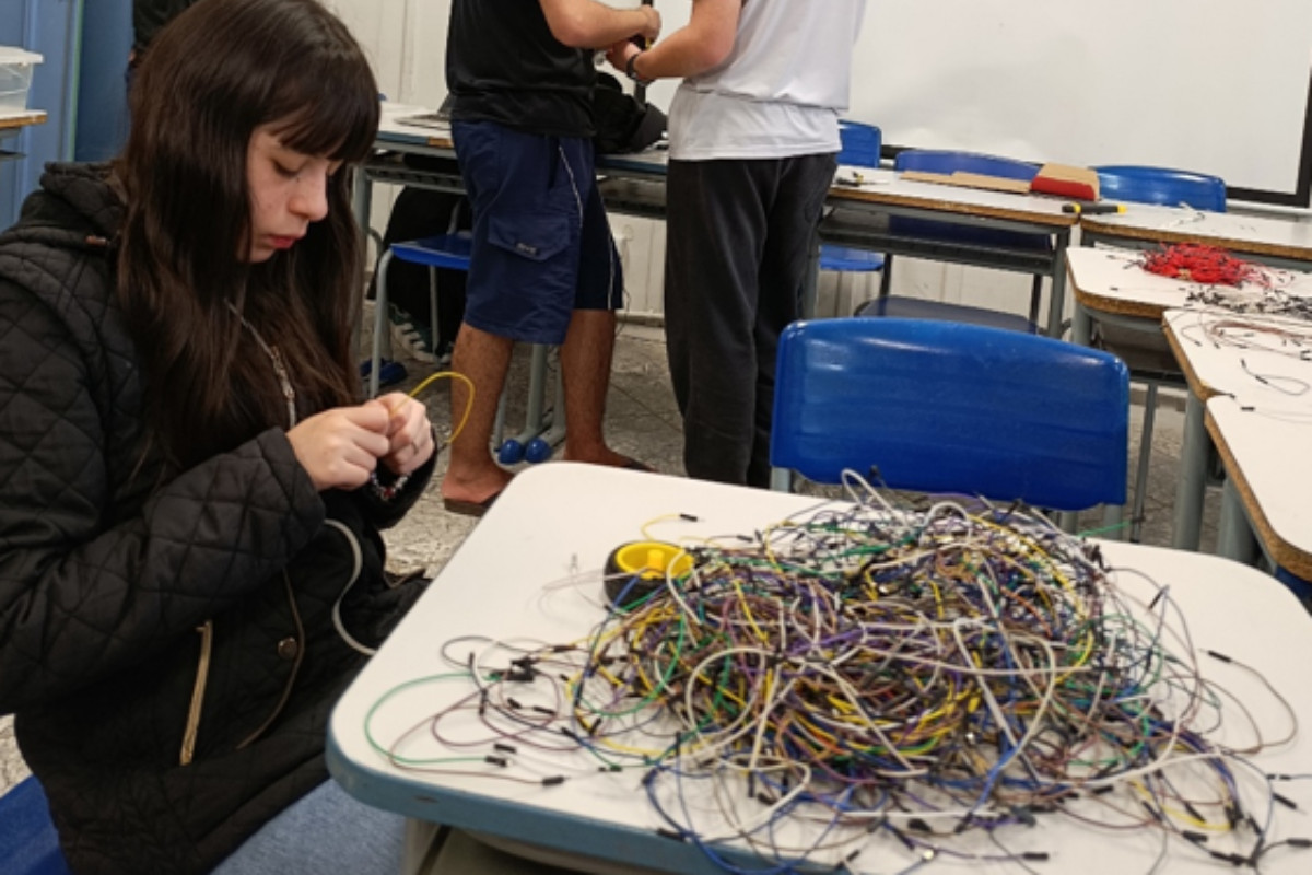 A imagem mostra um grupo de jovens em uma sala de aula, trabalhando em atividades práticas relacionadas à eletrônica. No primeiro plano, à esquerda, uma estudante está sentada, concentrada em manusear fios elétricos. Sobre a mesa à sua frente há um grande emaranhado de cabos coloridos, provavelmente utilizados em circuitos. Ao fundo, três rapazes estão em pé, colaborando em algum trabalho manual com fios ou componentes.