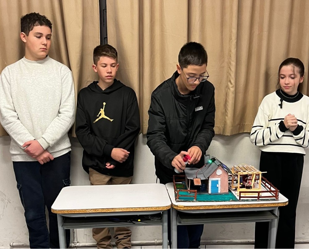 A imagem mostra quatro estudantes; dois deles, à esquerda, estão apenas observando, enquanto o terceiro, de óculos e casaco preto, manipula uma maquete apoiada sobre duas carteiras escolares. A maquete representa uma casa com telhado, cercas e estruturas anexas, sugerindo um projeto escolar de arquitetura, urbanismo, ciências ou tecnologia. À direita, uma aluna de blusa branca com listras pretas também observa atentamente a apresentação.