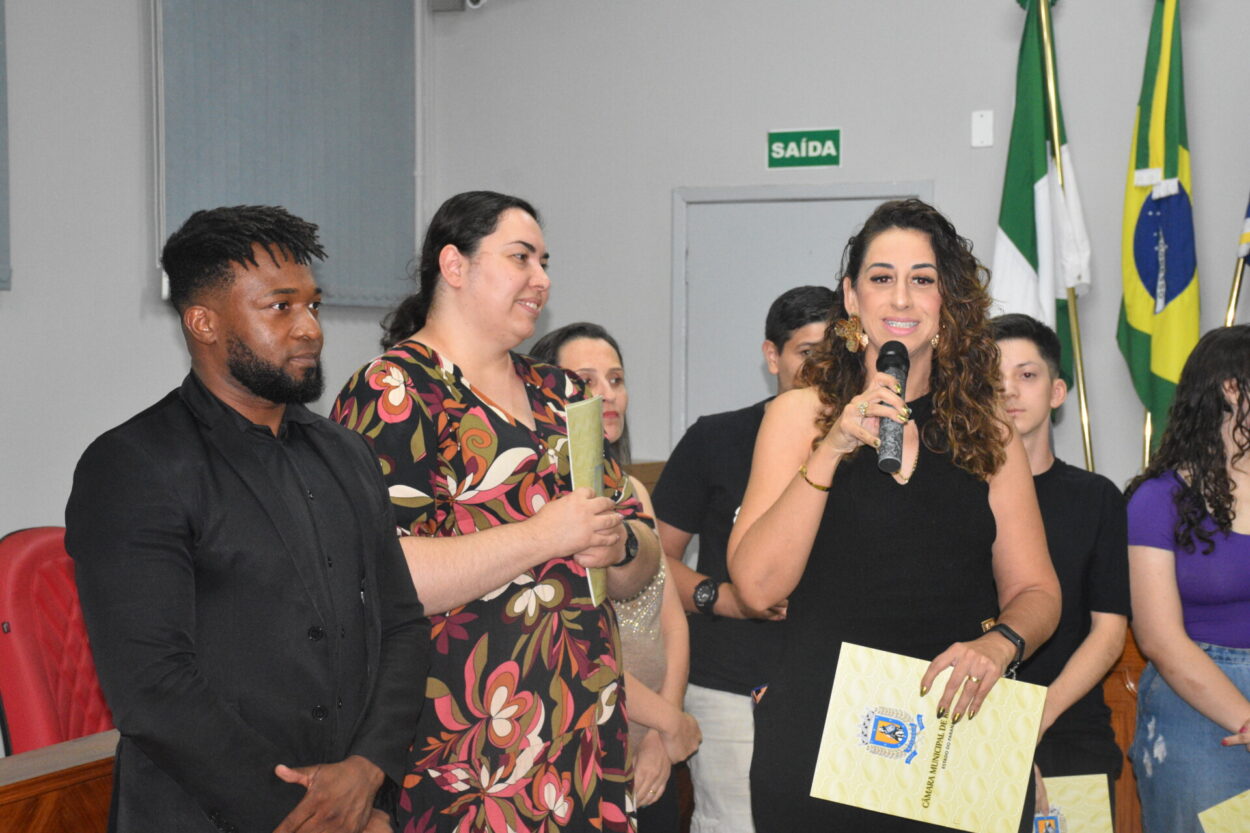 A foto mostra um grupo de pessoas dentro de uma câmara municipal. Em primeiro plano, uma mulher de cabelos cacheados e compridos fala ao microfone, segurando um certificado amarelo. Ela está vestida de preto e usa brincos dourados. Ao lado dela, estão um homem negro de terno escuro e uma mulher de cabelos presos, com um vestido estampado colorido, que segura outro certificado. Atrás deles, há mais pessoas em pé, algumas segurando papéis semelhantes.