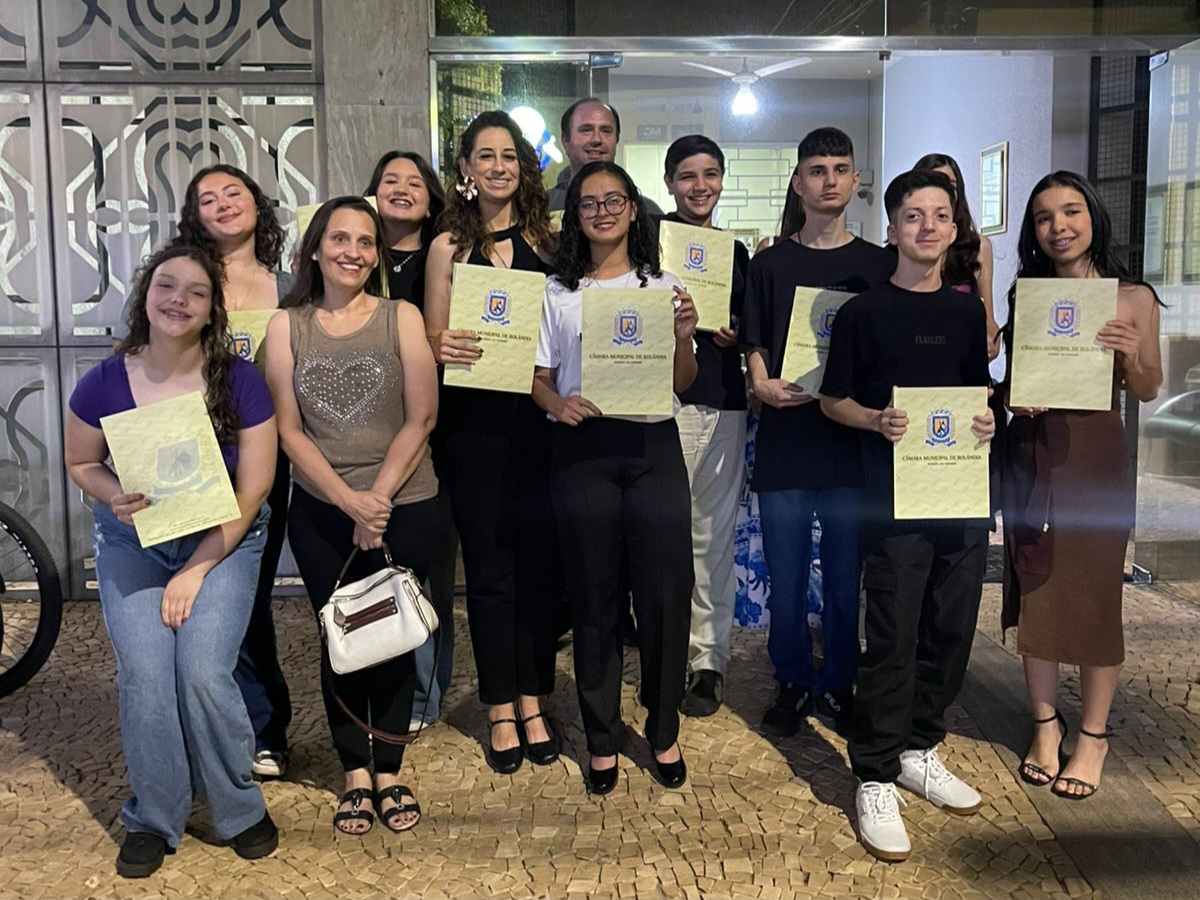 Esta foto foi tirada do lado de fora de um prédio, à noite. Um grupo de pessoas está posando para a foto, sorrindo e segurando certificados amarelos com o brasão da Câmara Municipal de Rolândia. Entre elas, há jovens e alguns adultos, segurando o documento. O clima é de comemoração e alegria.
