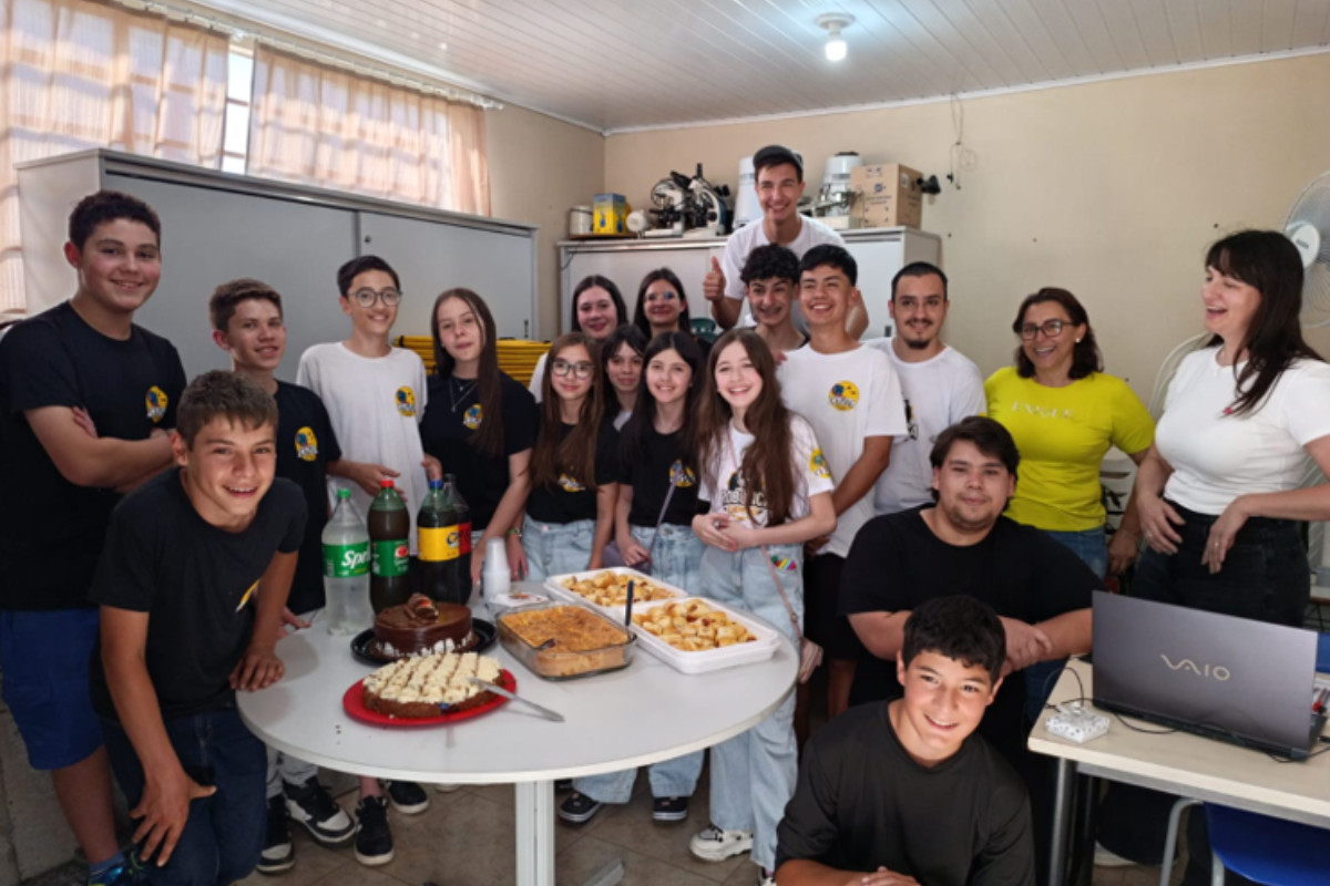A imagem mostra um grupo de pessoas reunidas em uma sala, celebrando uma confraternização. Na frente, há uma mesa redonda com comidas e bebidas. Algumas pessoas estão em pé atrás da mesa, enquanto outras estão sentadas ou agachadas à frente, todas sorrindo e olhando para a câmera. A maioria veste camisetas pretas ou brancas com o mesmo logotipo, indicando que fazem parte do projeto ligado à robótica (há a palavra ROBO em algumas camisetas). Duas mulheres, à direita, usam camisetas diferentes — uma amarela e outra branca, e parecem ser professoras ou responsáveis. No fundo da sala, há armários, prateleiras com equipamentos e caixas organizadas. Também se vê um notebook aberto sobre uma mesa lateral.