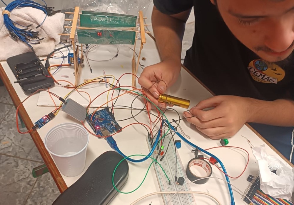A imagem mostra um jovem trabalhando em um projeto eletrônico sobre uma mesa. No centro da cena há diversos fios coloridos conectados a uma protoboard e a uma placa Arduino, indicando uma atividade de programação e montagem de circuito. Também aparecem outros materiais e componentes, usados para programar ou monitorar o sistema. Outros dois alunos estão ajudando e observando. O ambiente transmite a ideia de um trabalho prático de robótica ou eletrônica aplicada, combinando programação, montagem e testes experimentais.
