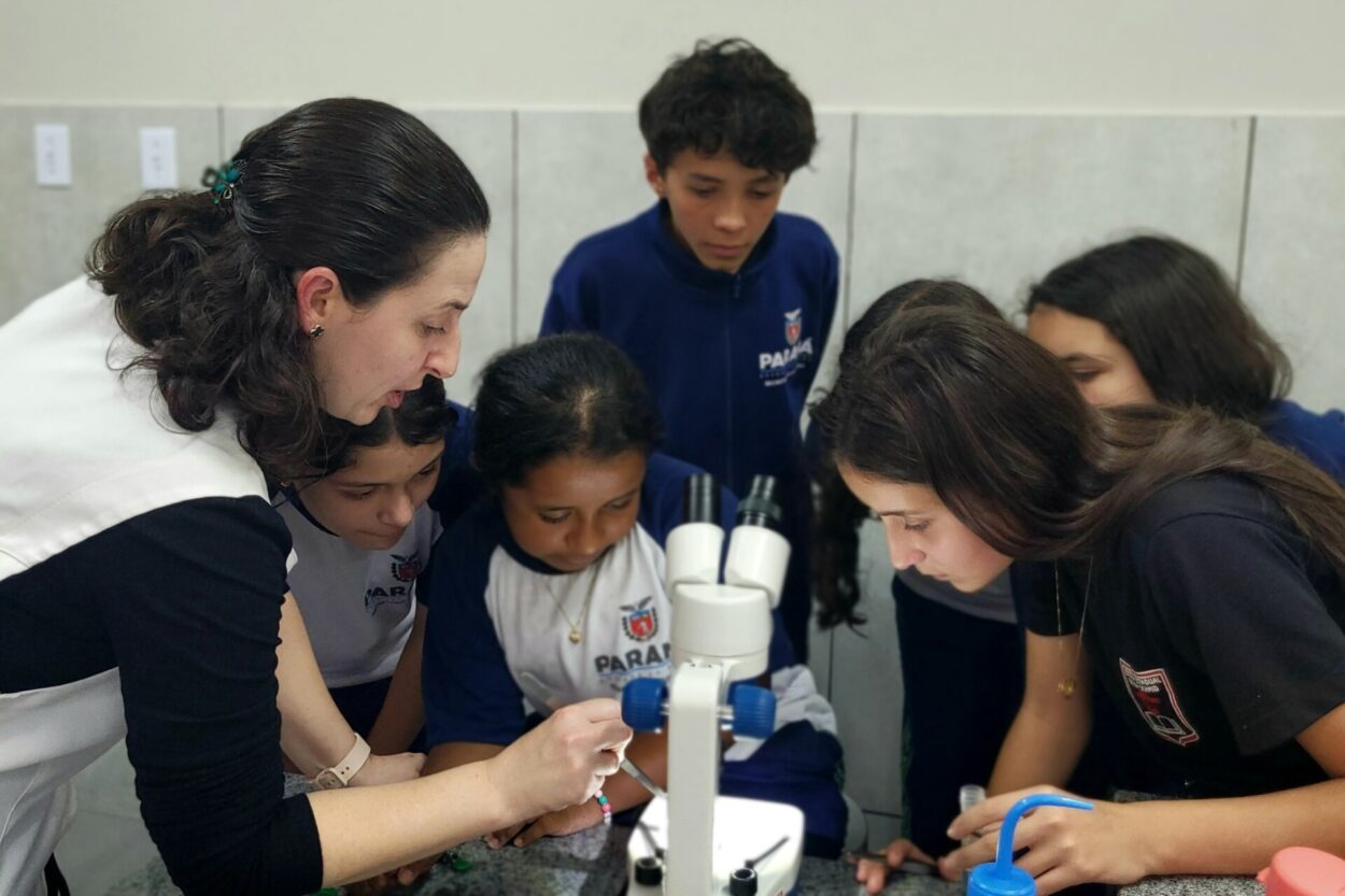 Um grupo de seis estudantes e uma professora participa de uma atividade em um laboratório escolar. À esquerda, a professora, de colete claro e blusa preta, orienta uma aluna que ajusta o foco de uma lupa binocular branca posicionada sobre a mesa. Ao redor, quatro estudantes observam atentos a cena, vestindo camisetas do uniforme azul e branco com o brasão do Paraná. À direita, uma estudante de camiseta preta acompanha de perto a análise feita no equipamento. Sobre a bancada de granito estão tubos de ensaio transparentes e um frasco azul de plástico. Ao fundo, a parede revestida de azulejos brancos completam o ambiente da atividade científica.