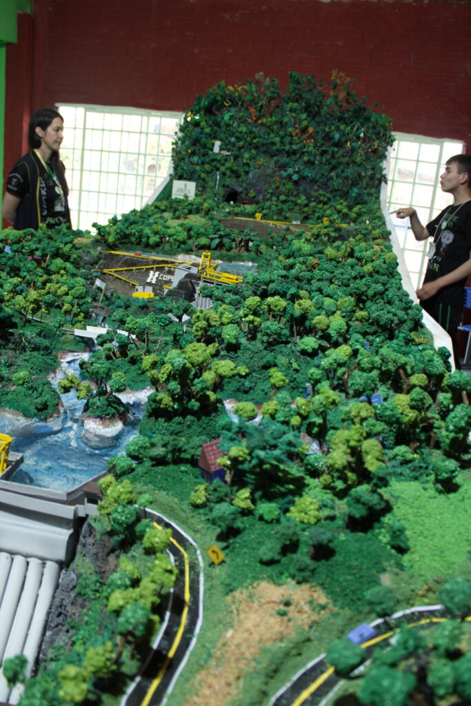 A foto mostra a maquete do clube Somos Fãs de Lavoisier, com mais de cinco metros de comprimento, ela representa o Rio Iguaçu e uma curiosidade é que a água realmente corre pela maquete. Na foto, um clubista e uma professora estão ao lado da maquete, que tem um bom ângulo de inclinação. No modelo, é possível observar pequenas árvores, casinhas, estradas, mini-hidrelétricas e é claro, o rio.