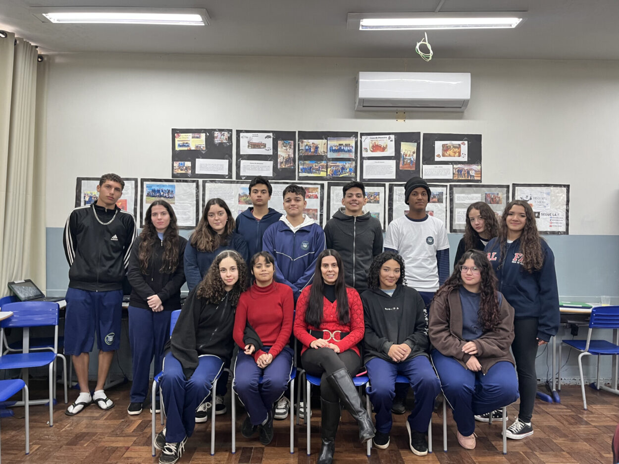 A foto é do clube de ciências ‘História e Mídia’ do Colégio Estadual Tomaz Edison de Andrade Vieira de Maringá. A imagem mostra um grupo de estudantes posando para uma foto dentro de uma sala de aula. O grupo é formado por 14 alunos e uma adulta, a professora. Eles estão dispostos em duas fileiras: a primeira sentada e a segunda em pé. Atrás deles há um mural com cartazes e trabalhos escolares expostos na parede. O ambiente é uma sala de aula com carteiras azuis, paredes, piso de madeira e iluminação de teto fluorescente.