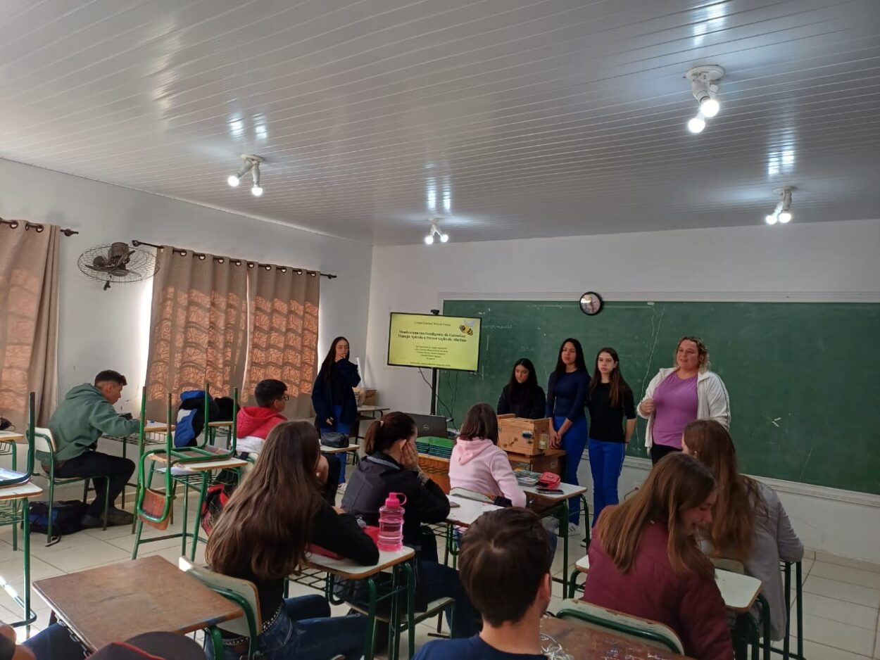 A foto mostra o grupo de estudantes reunidos em torno de uma mesa dentro de uma sala de aula, observando atentamente. Na mesa, há um o protótipo da colmeia inteligente, com fios e uma pequena tela ou display aceso. Ao fundo, há um quadro negro com escrita parcialmente apagada e um monitor ligado mostrando a tela inicial de um computador. A atmosfera sugere um momento de demonstração ou experimentação prática, com engajamento dos participantes.