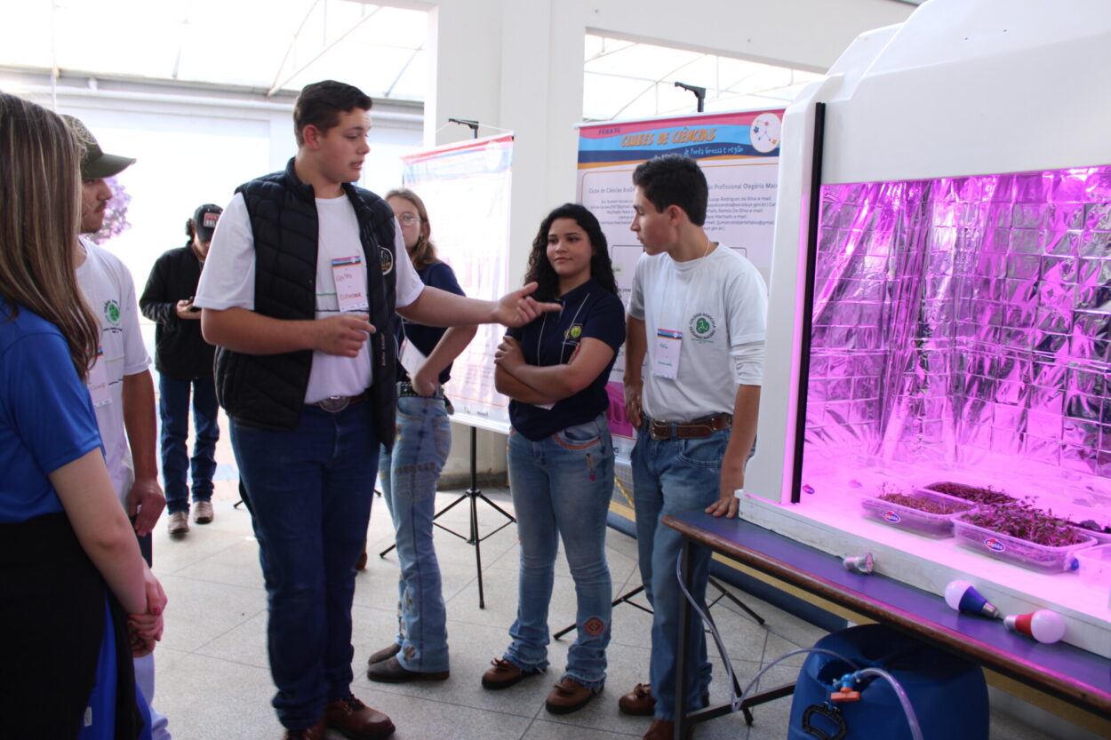 A foto foi tirada em um ambiente fechado, mas com forte iluminação natural, em primeiro plano e ao centro, um clubista do ecoindoor aponta para o protótipo de estrutura de cultivo indoor. O protótipo é uma grande caixa branca com iluminação rosa característica, em seu interior, um revestimento espelhado e bandejas com brotinhos. Na foto, vemos ainda mais clubistas observando seu colega apresentar e visitantes da feira interessados no trabalho, e banners no fundo