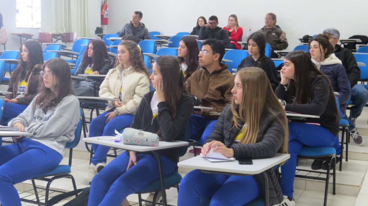 A imagem mostra uma sala de aula com estudantes sentados em carteiras, atentos a uma atividade. A maioria veste calças azuis e casacos, indicando uniforme escolar. Em primeiro plano, algumas alunas observam para frente, enquanto fazem anotações. Ao fundo, há mais estudantes e alguns adultos também acompanhando a atividade. As carteiras são brancas e azuis, e o ambiente é iluminado pela luz natural que entra pelas janelas.