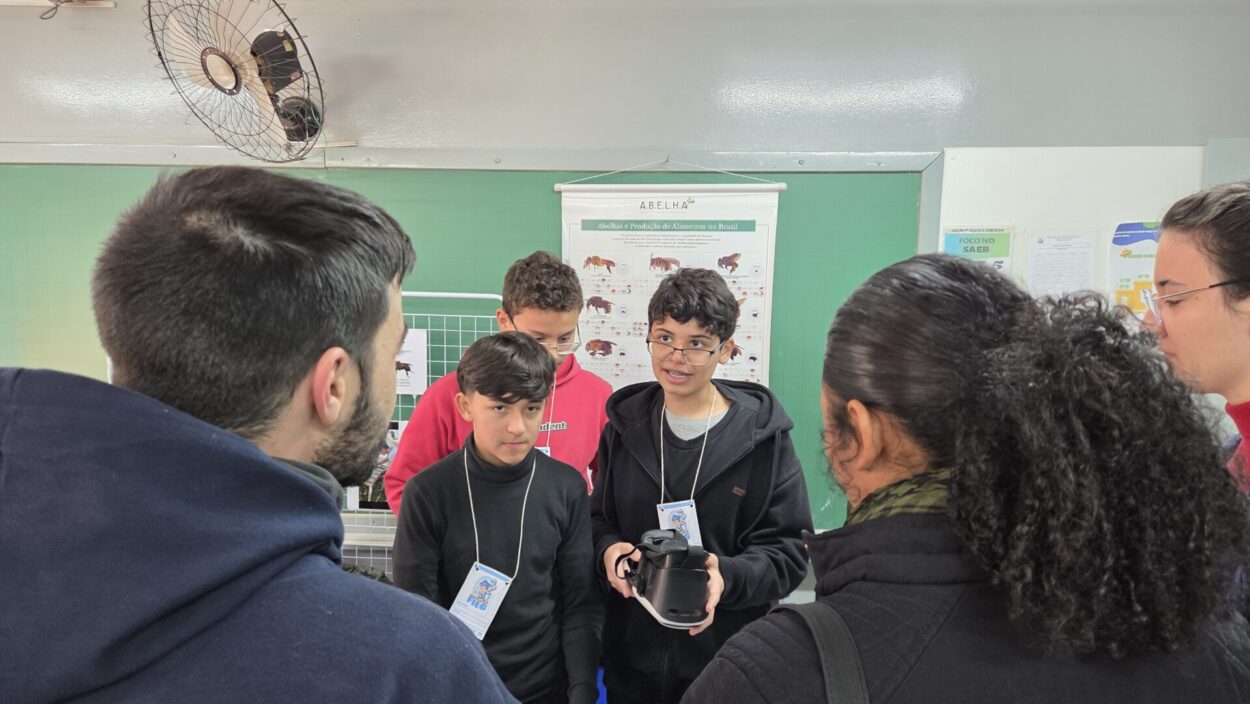 Na imagem, alguns estudantes apresentam um projeto em uma sala de aula. Dois deles estão à frente, um segurando um equipamento de realidade virtual, enquanto outras pessoas observam atentamente. Ao fundo, há cartazes educativos fixados na parede verde.