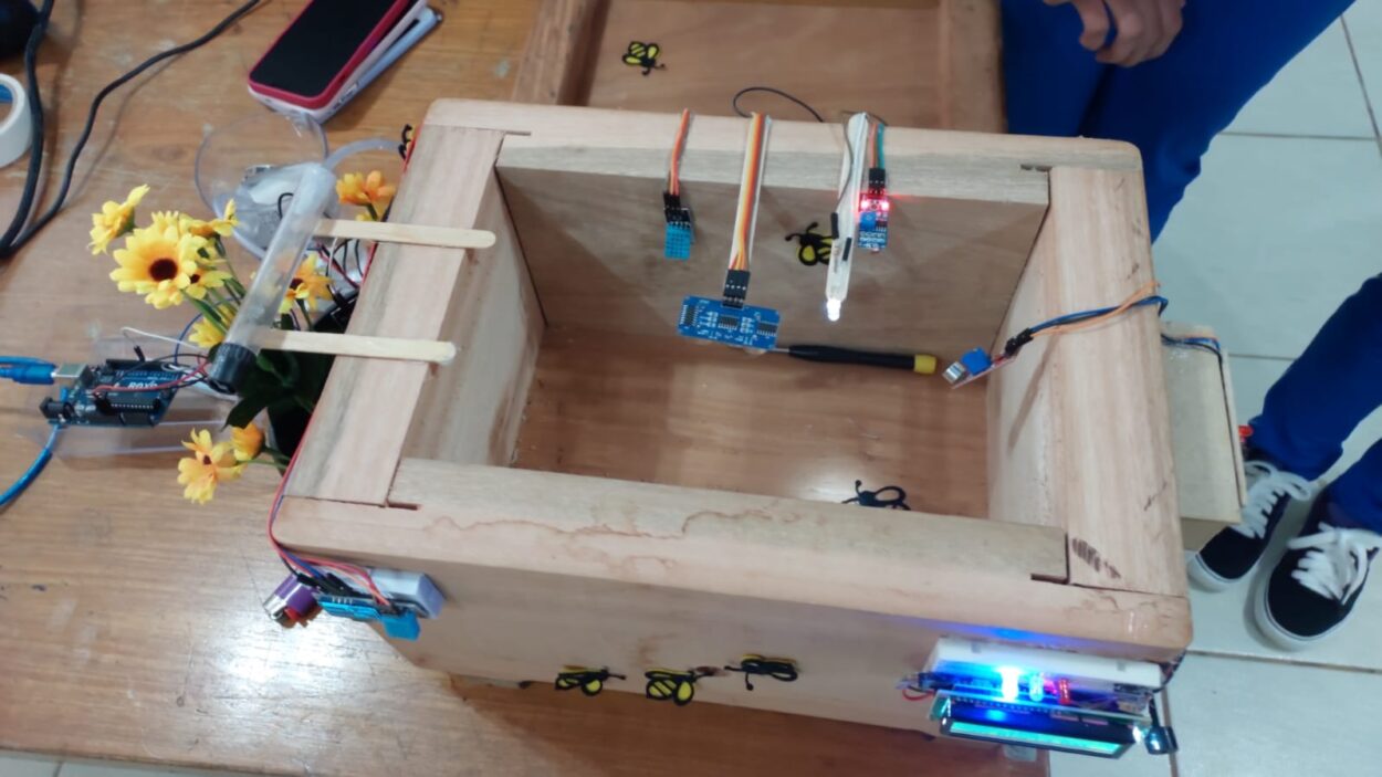 A imagem mostra um protótipo de colmeia inteligente, feito principalmente de madeira, colocado sobre uma mesa de madeira clara. No interior e nas laterais da colmeia, estão conectados diversos sensores eletrônicos, com fios coloridos ligados a um microcontrolador e a pequenos módulos de monitoramento. Na parte externa, há pequenas luzes indicadoras acesas em azul e vermelho. No canto esquerdo da colmeia, há um arranjo de flores artificiais amarelas, possivelmente simulando um ambiente natural para as abelhas. Alguns adesivos de abelhas estão colados nas superfícies da madeira.