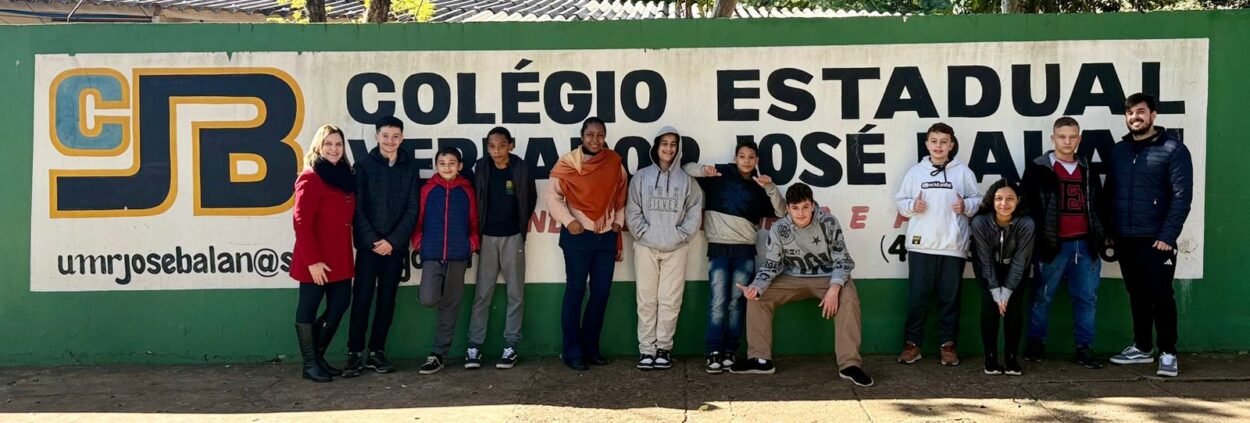 A foto é dos alunos do Clube Dengue Bot do Colégio Estadual Vereador José Balan de Umuarama com projetos finalistas no FENAAH/S. A imagem mostra um grupo de pessoas posando em frente a um muro verde com uma placa do Colégio Estadual Vereador José Balan. Ao fundo, há uma grande pintura com o logotipo "CJB" em destaque, seguido do nome completo da escola em letras grandes e pretas. O grupo é composto por 13 pessoas, incluindo jovens e dois adultos. À esquerda da imagem, uma mulher de casaco vermelho e cachecol preto está sorrindo, ao lado de vários estudantes. À direita, outro adulto, de barba e vestindo jaqueta escura, também posa ao lado dos alunos. Todos estão agasalhados, sugerindo que a foto foi tirada em um dia frio. O clima é de descontração e união.