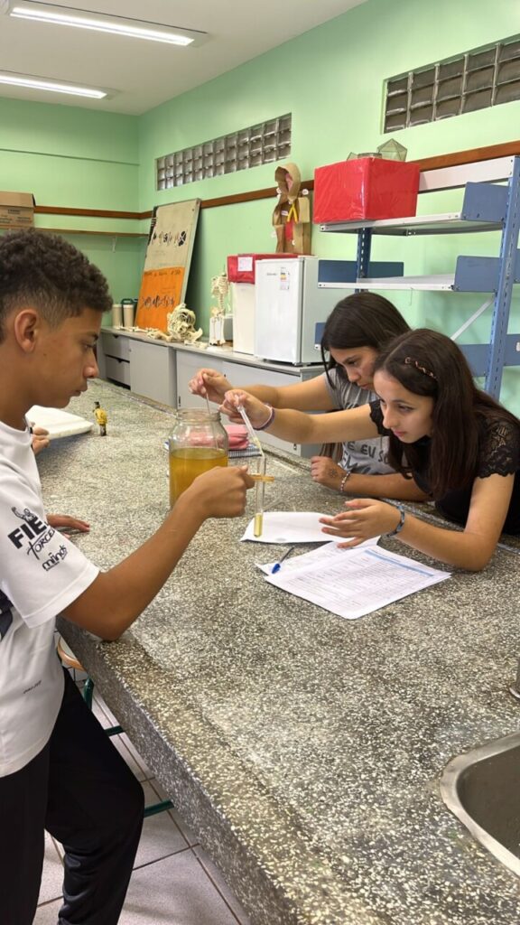 A foto mostra clubistas em laboratório, em uma bancada de mármore, eles fazem testes com óleo de cozinha. Esse está armazenado em um grande pote de vidro, sendo colocado em um tubo de ensaio.