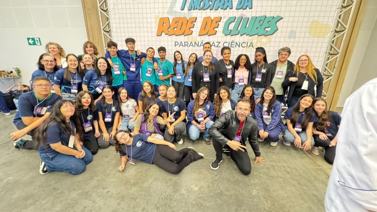 Na foto apresentada, é possível ver no fundo da imagem atrás dos alunos e professores, em destaque, a I Mostra da Rede de Clubes Paraná FAZ Ciência, todos celebram a participação no dia da feira, depois de um dia muito produtivo.