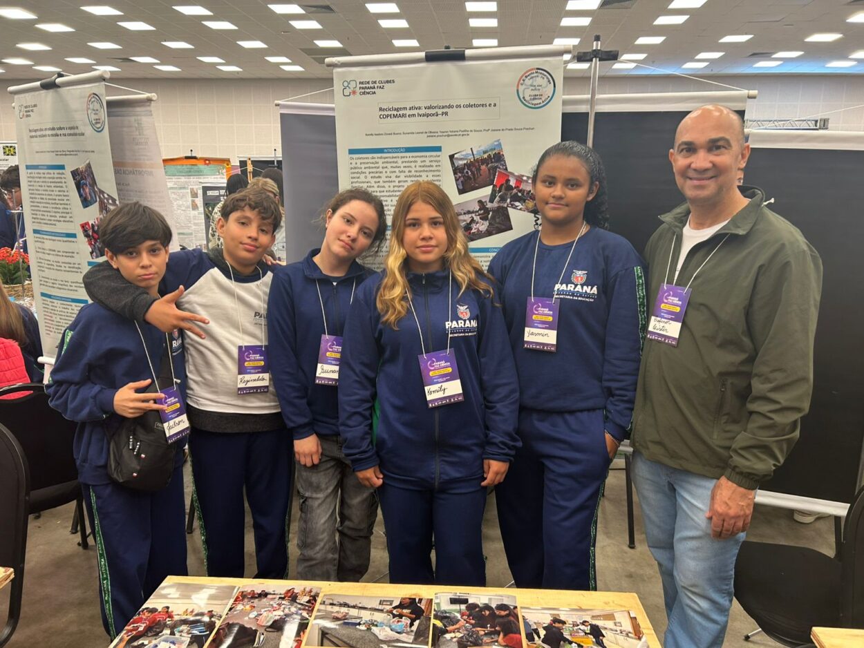 A foto é do clube de ciências ‘Reciclagem Ativa’ do Colégio Estadual Bento Mossurunga de Ivaiporã. A imagem mostra um grupo formado por cinco estudantes e o professor, posando juntos em um evento científico. Eles estão em um ambiente de exposição, com pôsteres científicos ao fundo, que trazem textos e imagens relacionados a projetos escolares. Os estudantes vestem uniformes da rede estadual do Paraná. Cada um usa um crachá roxo pendurado no pescoço, indicando participação oficial no evento. À frente do grupo há uma mesa com várias fotos impressas que parecem registrar atividades práticas ou etapas do projeto apresentado. O ambiente é bem iluminado, com várias pessoas e estandes ao fundo, sugerindo uma feira de ciências.
