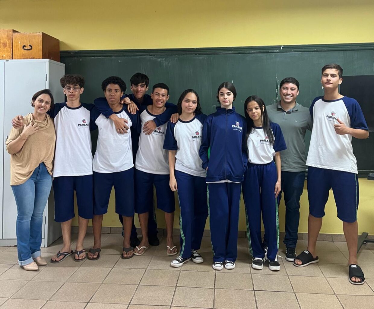 Na primeira fotografia é possível ver um grupo de pessoas posando juntas dentro de uma sala de aula. Ao todo, são dez pessoas: oito jovens e dois adultos. Eles estão posicionados lado a lado. O ambiente é típico de sala de aula, com paredes amarelas, quadro verde ao fundo e piso de cerâmica. A segunda imagem é do Clube Ilha da Ciência. A imagem mostra um grupo de estudantes e duas mulheres posando em um ambiente ao ar livre, com árvores, arbustos e canteiros de horta ao fundo. O grupo está sorridente, alguns em pé e outros agachados na frente. Na terceira fotografia tem um grupo de estudantes reunidos em um ambiente escolar ao ar livre, em frente a um jardim vertical com diversas plantas verdes. Eles estão distribuídos em pé e agachados, posando para a foto. A quarta imagem mostra um grupo de estudantes e um adulto reunidos em um espaço ao ar livre, posando para uma foto em frente a uma grande escultura de uma figura humana com os braços abertos. O grupo está distribuído sobre um pequeno morro coberto de grama, alguns sentados e outros em pé. A quinta imagem é do Clube Experimentando a Ciência: Descobrindo cores e novos sabores. A imagem mostra um grupo de estudantes e professores posando para uma foto ao ar livre, em frente a uma placa de madeira com o desenho de uma abelha e a palavra “Meliponário”. Estão sorridentes e organizados ao redor da placa. Na sexta imagem mostra um grupo grande de pessoas, formado por adolescentes e um adulto, posando para uma foto em uma sala. Eles estão organizados em duas fileiras: alguns sentados ou agachados na frente. A sala tem paredes claras, ventiladores fixados na parede e um quadro branco ao fundo. À esquerda, há uma mesa com uma toalha vermelha e alguns objetos decorativos para um evento escolar. A última fotografia tem um grupo de estudantes e alguns adultos reunidos para uma foto em um ambiente escolar externo. Eles estão em frente a uma parede branca com detalhes verdes e uma janela comprida com grades. O grupo está posicionado em duas fileiras, com alguns agachados na frente e outros em pé atrás, todos sorrindo.