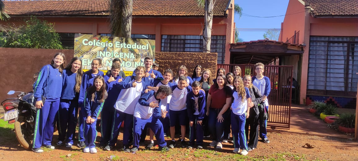 A primeira imagem mostra um grupo de estudantes posando para uma foto em frente a uma escola. O grupo é composto por cerca de vinte jovens e alguns adultos, todos sorrindo. Eles estão ao ar livre, em um local ensolarado, com solo de terra vermelha e vegetação ao redor. Ao fundo, vê-se o prédio da escola, de paredes alaranjadas e telhado de cerâmica, com janelas amplas e uma entrada com portão de ferro. Na segunda imagem aparece um jovem, vestido com uniforme escolar azul e branco, praticando arco e flecha em um campo gramado. Ele segura o arco esticado, mirando para frente, concentrado no alvo. À esquerda, um homem indígena com cocar de penas vermelhas observa atentamente o aluno, aparentando orientá-lo na atividade. Outros dois homens estão mais ao fundo — um em pé, vestindo camiseta cinza com detalhes coloridos, e dois sentados na grama, observando a prática. O cenário é ensolarado, com gramado verde, árvores e um prédio de paredes alaranjadas e janelas amplas ao fundo. A última fotografia tem um grupo de pessoas participando de uma atividade ao ar livre em um campo gramado. Elas estão alinhadas lado a lado, segurando arcos e flechas, aparentemente se preparando para praticar ou competir em uma prova de tiro com arco. Entre os participantes, há jovens e adultos. Um homem, à frente do grupo, parece estar orientando ou demonstrando a técnica do disparo. Ao fundo, há árvores altas, uma cerca de madeira e algumas tendas ou coberturas improvisadas, sob as quais outras pessoas estão sentadas.