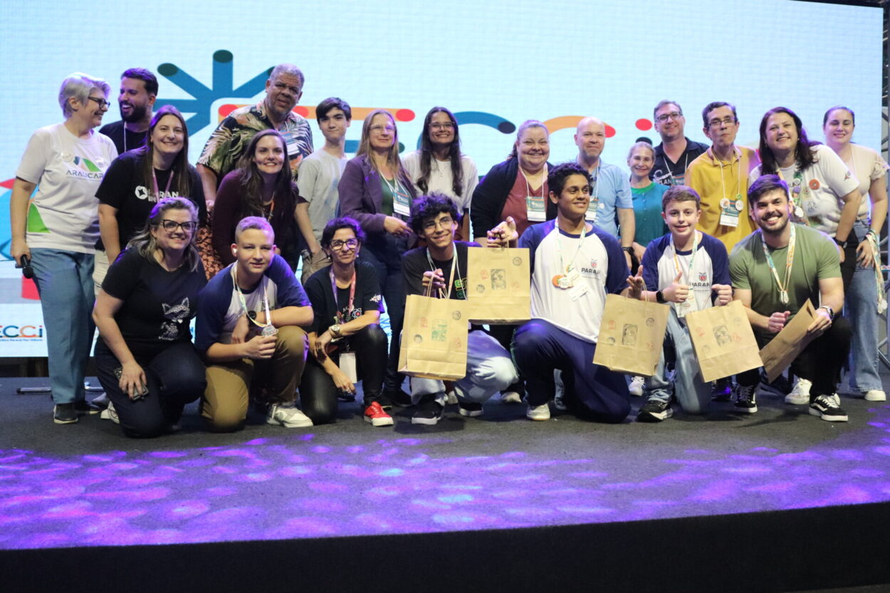 A primeira imagem é do Clube de Ciências Tenda Cultura Viva, do Colégio Estadual Professora Hilda Trautwein Kamal. Na foto há um grupo de pessoas reunidas sobre um palco, sorrindo e posando para a foto durante um evento. Ao fundo, há um telão sugerindo um encontro educacional. Os participantes estão lado a lado, segurando certificados, sacolas de papel e prêmios. Uma pessoa ao centro segura uma grande caixa de uma caixa de som JBL. Todos usam crachás de identificação e vestem roupas casuais. Na segunda fotografia vemos o Dengue Bot, do Colégio Estadual Vereador José Balan. Um grupo grande de pessoas reunidas sobre um palco, sorrindo para uma foto coletiva. Ao fundo, há um telão iluminado com elementos gráficos coloridos, sugerindo que o evento é de caráter educacional. Na parte da frente, alguns jovens seguram sacolas de papel e usam crachás de identificação e camisetas com o logotipo do Paraná. O Paisagismo Sustentável da Escola Estadual Manuel Bandeira, de Alto Piquiri, é a terceira foto. A imagem mostra um grupo numeroso de pessoas reunidas sobre um palco, posando para uma foto durante um evento educacional. Ao fundo, há um telão iluminado com elementos gráficos, reforçando o clima festivo e institucional. Na parte da frente, alguns jovens e adultos exibem certificados, troféus e prêmios. A quarta foto é do Clube Green Lab do Colégio Estadual Almirante Tamandaré, de Cruzeiro do Oeste. Na imagem é possível ver pessoas posando juntas em um palco. Há cerca de quinze pessoas, entre adultos e jovens, algumas agachadas na frente e outras em pé atrás. Os jovens na frente seguram certificados e sacolas de papel. No fundo, há um telão grande com um logotipo colorido.