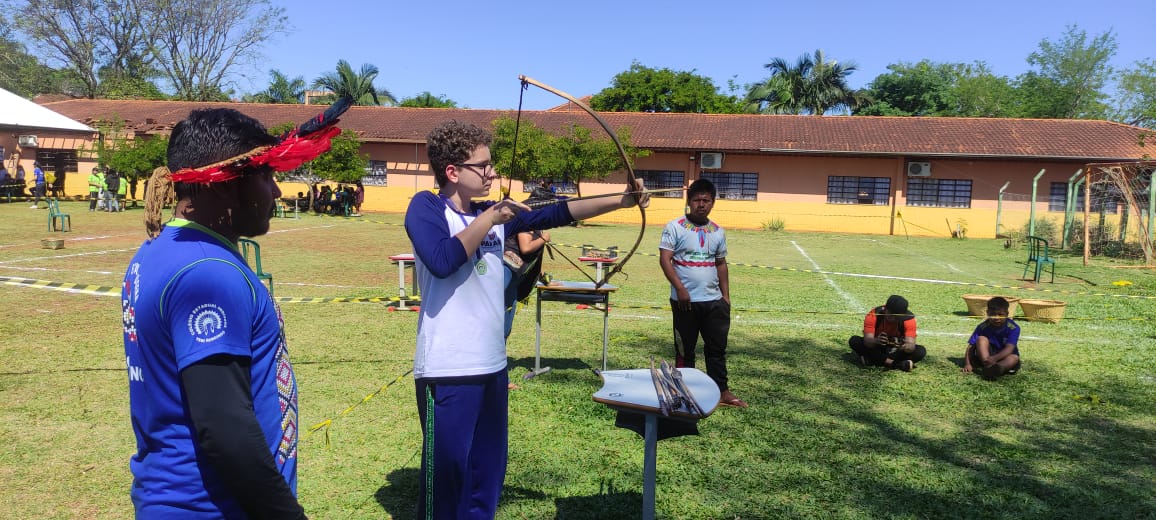 A primeira imagem mostra um grupo de estudantes posando para uma foto em frente a uma escola. O grupo é composto por cerca de vinte jovens e alguns adultos, todos sorrindo. Eles estão ao ar livre, em um local ensolarado, com solo de terra vermelha e vegetação ao redor. Ao fundo, vê-se o prédio da escola, de paredes alaranjadas e telhado de cerâmica, com janelas amplas e uma entrada com portão de ferro. Na segunda imagem aparece um jovem, vestido com uniforme escolar azul e branco, praticando arco e flecha em um campo gramado. Ele segura o arco esticado, mirando para frente, concentrado no alvo. À esquerda, um homem indígena com cocar de penas vermelhas observa atentamente o aluno, aparentando orientá-lo na atividade. Outros dois homens estão mais ao fundo — um em pé, vestindo camiseta cinza com detalhes coloridos, e dois sentados na grama, observando a prática. O cenário é ensolarado, com gramado verde, árvores e um prédio de paredes alaranjadas e janelas amplas ao fundo. A última fotografia tem um grupo de pessoas participando de uma atividade ao ar livre em um campo gramado. Elas estão alinhadas lado a lado, segurando arcos e flechas, aparentemente se preparando para praticar ou competir em uma prova de tiro com arco. Entre os participantes, há jovens e adultos. Um homem, à frente do grupo, parece estar orientando ou demonstrando a técnica do disparo. Ao fundo, há árvores altas, uma cerca de madeira e algumas tendas ou coberturas improvisadas, sob as quais outras pessoas estão sentadas.