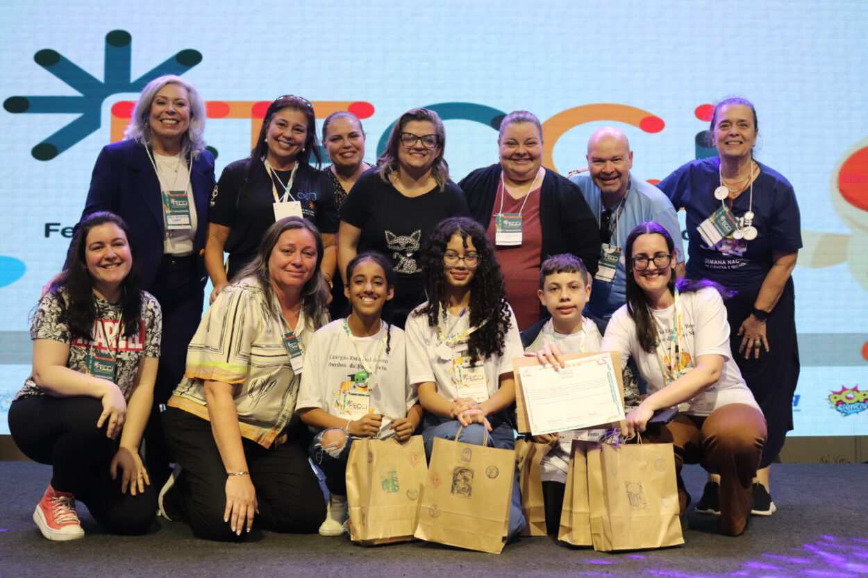 A foto mostra um grupo de treze pessoas posando juntas em um palco. Na frente, três crianças estão sentadas ou agachadas, segurando sacolas de papel e um certificado.Ao lado delas, duas mulheres adultas também estão agachadas, aproximando-se do grupo infantil com expressão alegre. Atrás, há oito adultos em pé, alinhados, todos sorrindo. O fundo é um painel grande e iluminado, exibindo um logotipo colorido e abstrato.