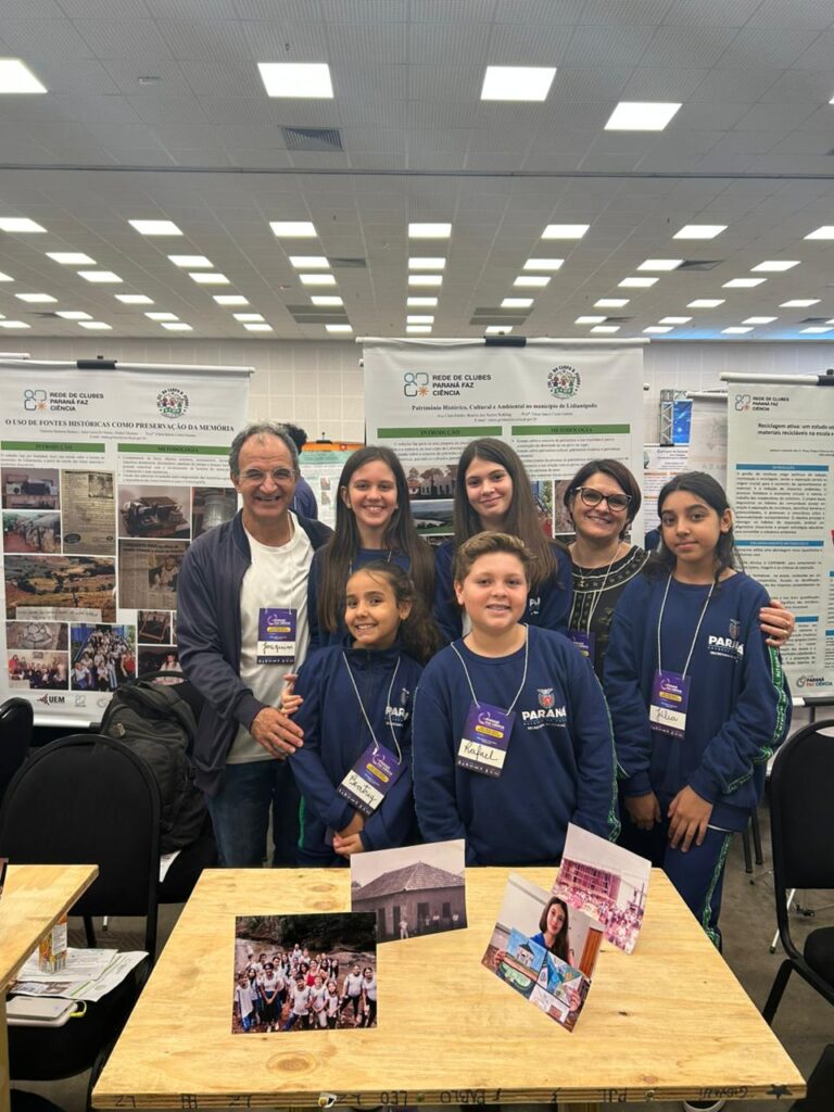 A primeira fotografia mostra um grupo formado por cinco estudantes e o professor, posando juntos em um evento científico. Eles estão em um ambiente de exposição, com pôsteres científicos ao fundo, que trazem textos e imagens relacionados a projetos escolares. Os estudantes vestem uniformes da rede estadual do Paraná. A segunda imagem é do Clube Show da Química. A imagem mostra um grupo de cinco pessoas, três estudantes e dois adultos, posando em um evento educacional. Eles estão em pé atrás de uma mesa de madeira com alguns materiais usados em experimentos, como garrafas plásticas com líquidos e copos descartáveis. O ambiente é um grande salão, bem iluminado, com outras pessoas, estandes e pôsteres ao fundo, reforçando o clima de evento científico. Na terceira fotografia tem um grupo formado por seis pessoas — quatro estudantes e dois adultos — posando juntos em um ambiente de feira científica. É o clube “Entre o passado e o presente, o que fica é a memória da gente”. Eles estão de pé atrás de uma mesa de madeira onde há várias fotos impressas que ilustram as atividades do projeto, incluindo imagens de grupos de alunos e de locais históricos. Ao fundo, há pôsteres científicos com o logotipo da Rede de Clubes Paraná Faz Ciência. A quarta imagem mostra quatro pessoas em um evento educacional, posando ao lado de uma mesa com materiais de um projeto sobre compostagem e reciclagem orgânica. Na frente, um jovem segura um balde verde identificado com o logotipo “C.C.H.E” e a imagem de uma minhoca, enquanto outro rapaz, ao lado, segura um copo transparente com um líquido escuro. No centro da mesa há um cesto com minhocas californianas e outro balde verde com ilustrações educativas relacionadas ao ciclo da compostagem. Atrás deles, uma moça e um homem adulto completam o grupo, todos usando crachás roxos de identificação do evento. A quinta imagem é do Clube Natureza em Perspectiva. A imagem mostra um grupo de estudantes posando para uma foto em um ambiente escolar ao ar livre. Ao todo, são cerca de vinte alunos, acompanhados por duas adultas, formando uma fileira de pé e outra agachada à frente. O grupo está em frente a um muro colorido e decorado com desenhos alegres, incluindo um sol, nuvens, flores e elementos da natureza.