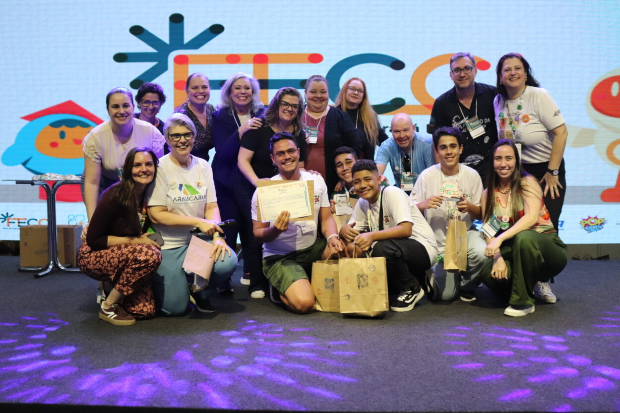 A foto é do Clube de Ciências ‘Robótica e Sustentabilidade, Solidariedade em Ação’ da Escola Estadual Professor Giampero Monacci de Itambé. A imagem mostra um grupo de pessoas reunidas sobre um palco, sorrindo e posando para a foto em um evento. Ao fundo, há um telão com logotipos coloridos e figuras ilustradas. Na parte da frente, três jovens e um homem estão ajoelhados, segurando certificados e sacolas de papel. Atrás deles, há várias pessoas adultas também sorrindo e demonstrando alegria pela conquista. Alguns participantes vestem camisetas brancas com o logotipo da “Araucária”, enquanto outros usam crachás e roupas casuais.