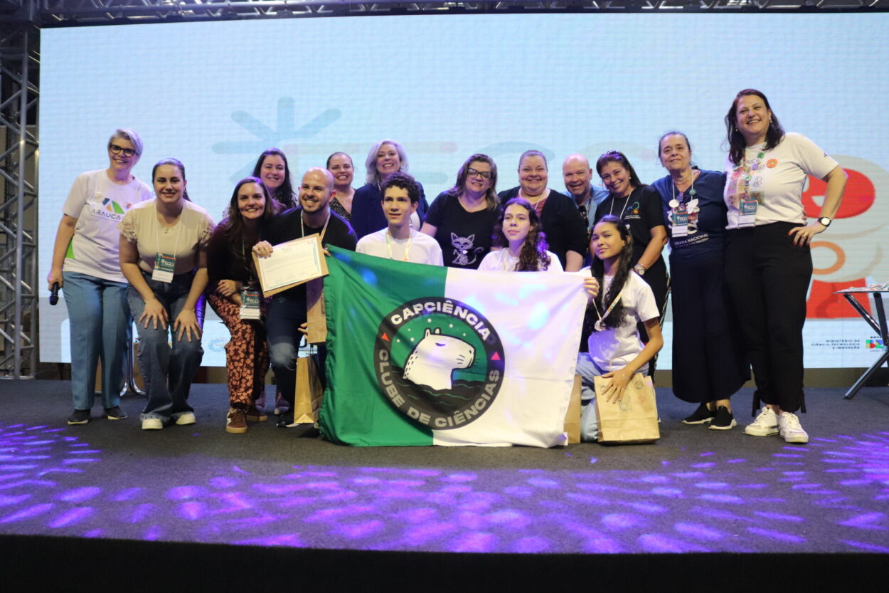 A primeira imagem é do Clube de Ciências ‘Robótica e Sustentabilidade, Solidariedade em Ação’ da Escola Estadual Professor Giampero Monacci de Itambé. A imagem mostra um grupo de pessoas reunidas sobre um palco, sorrindo e posando para a foto em um evento. Ao fundo, há um telão com logotipos coloridos e figuras ilustradas. A segunda imagem é do Clube Capivara’s Tech do Colégio Estadual Brasílio Itiberê. A imagem mostra um grupo de pessoas posando sorridentes sobre um palco, durante um evento. Ao fundo, há um telão com logotipos coloridos e desenhos estilizados, indicando um ambiente educacional. Na parte da frente, algumas pessoas estão agachadas, segurando certificados e sacolas de papel. Atrás delas, há outras pessoas, também sorrindo e comemorando. Na terceira fotografia do Clube CAPCiência do Colégio de Aplicação Pedagógica da UEM há um grupo de pessoas reunidas sobre um palco, sorrindo para a foto durante um evento. No centro, alguns participantes seguram uma bandeira verde e branca com o logotipo do “Capciência – Clube de Ciências”, que traz a ilustração de uma capivara. Atrás deles, há um grupo de adultos, todos usando crachás de identificação.