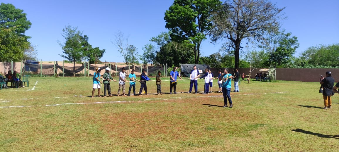 A primeira imagem mostra um grupo de estudantes posando para uma foto em frente a uma escola. O grupo é composto por cerca de vinte jovens e alguns adultos, todos sorrindo. Eles estão ao ar livre, em um local ensolarado, com solo de terra vermelha e vegetação ao redor. Ao fundo, vê-se o prédio da escola, de paredes alaranjadas e telhado de cerâmica, com janelas amplas e uma entrada com portão de ferro. Na segunda imagem aparece um jovem, vestido com uniforme escolar azul e branco, praticando arco e flecha em um campo gramado. Ele segura o arco esticado, mirando para frente, concentrado no alvo. À esquerda, um homem indígena com cocar de penas vermelhas observa atentamente o aluno, aparentando orientá-lo na atividade. Outros dois homens estão mais ao fundo — um em pé, vestindo camiseta cinza com detalhes coloridos, e dois sentados na grama, observando a prática. O cenário é ensolarado, com gramado verde, árvores e um prédio de paredes alaranjadas e janelas amplas ao fundo. A última fotografia tem um grupo de pessoas participando de uma atividade ao ar livre em um campo gramado. Elas estão alinhadas lado a lado, segurando arcos e flechas, aparentemente se preparando para praticar ou competir em uma prova de tiro com arco. Entre os participantes, há jovens e adultos. Um homem, à frente do grupo, parece estar orientando ou demonstrando a técnica do disparo. Ao fundo, há árvores altas, uma cerca de madeira e algumas tendas ou coberturas improvisadas, sob as quais outras pessoas estão sentadas.