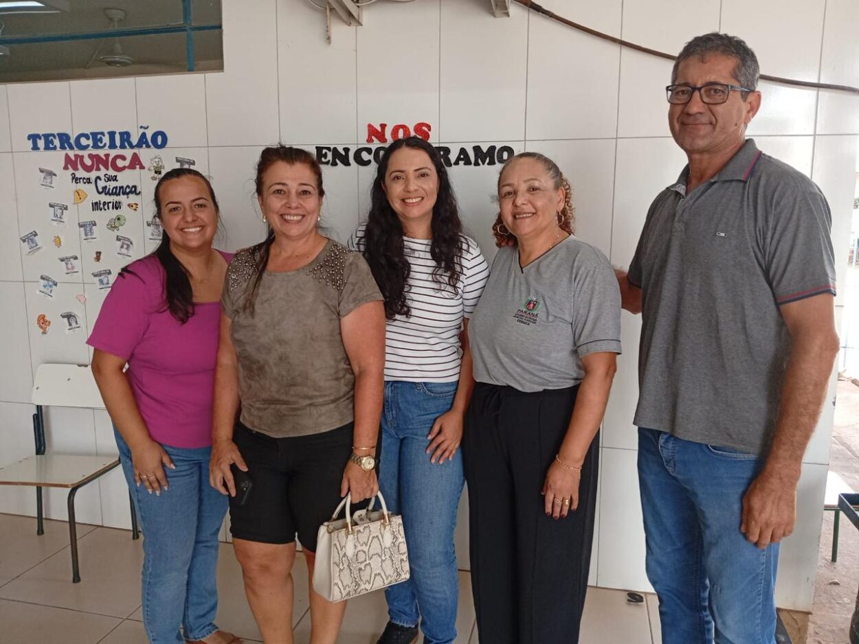 imagem mostra cinco pessoas em pé, lado a lado, sorrindo para a câmera em um ambiente interno, provavelmente escolar. Ao fundo, há um mural com frases coloridas e desenhos. As paredes são revestidas com azulejos brancos. As pessoas aparentam estar em um momento de confraternização ou registro de uma visita, demonstrando simpatia e cordialidade.