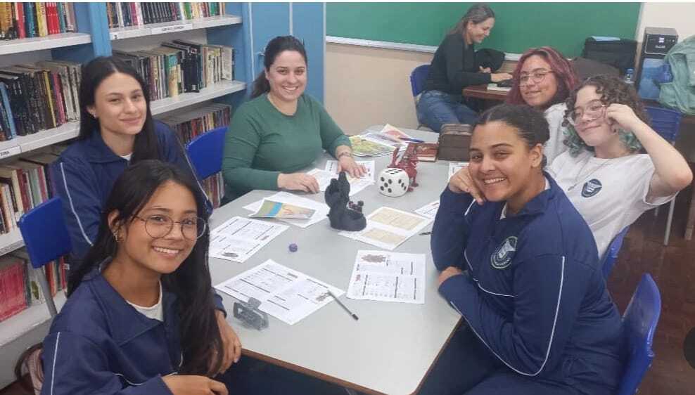 A foto mostra a equipe do clube, sentadas e posando para a câmera. Ao fundo, há estandes de livros, indicando uma biblioteca.